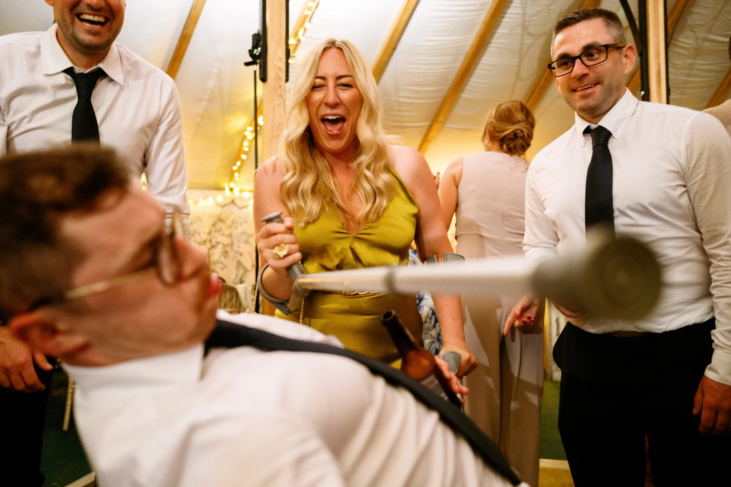 Group of people at a celebration or wedding, with one woman in a yellow dress laughing and holding a microphone near a seated man with glasses.