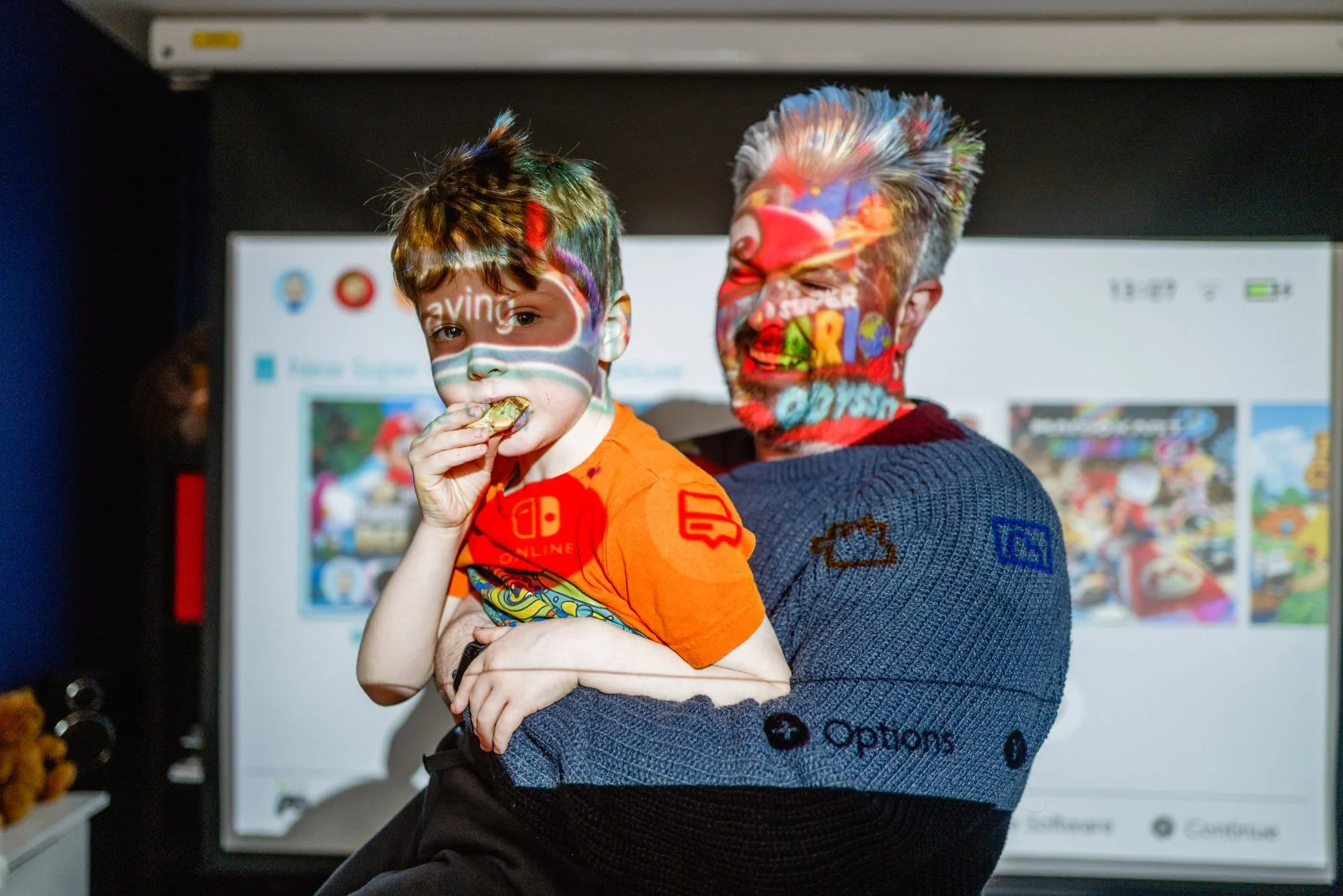 A man with gray hair and a beard holds a young boy in front of a television screen, both with colorful projected graphics on their faces and clothing. The boy is eating a snack and wearing an orange shirt.