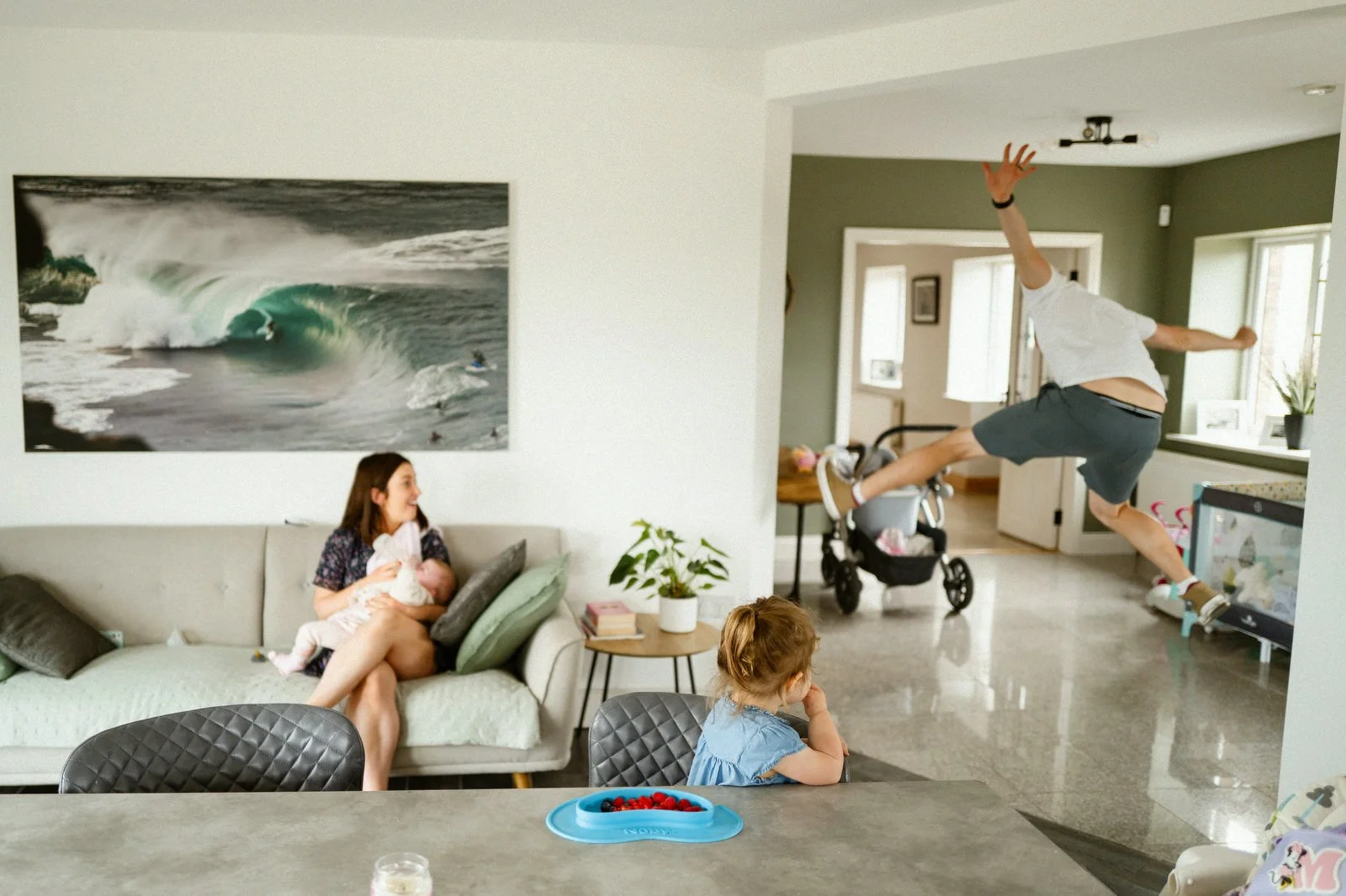 A family in their living room, with one man jumping in the air near the doorway, two young girls sitting at the table, one holding a baby, and a large ocean wave picture on the wall.