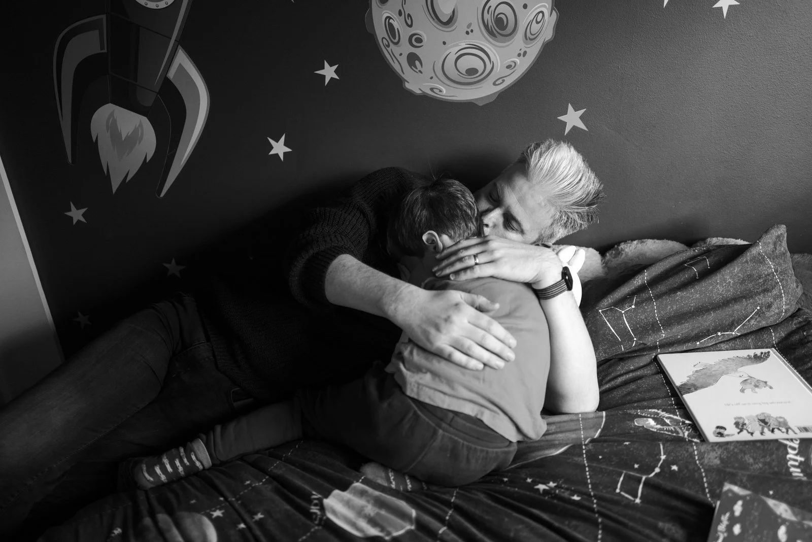 A person with short, bleached hair is lying on a bed, hugging a young boy. The background features a space-themed wall with stars and a rocket illustration, as well as a moon drawing. A book with animal illustrations is on the bed.