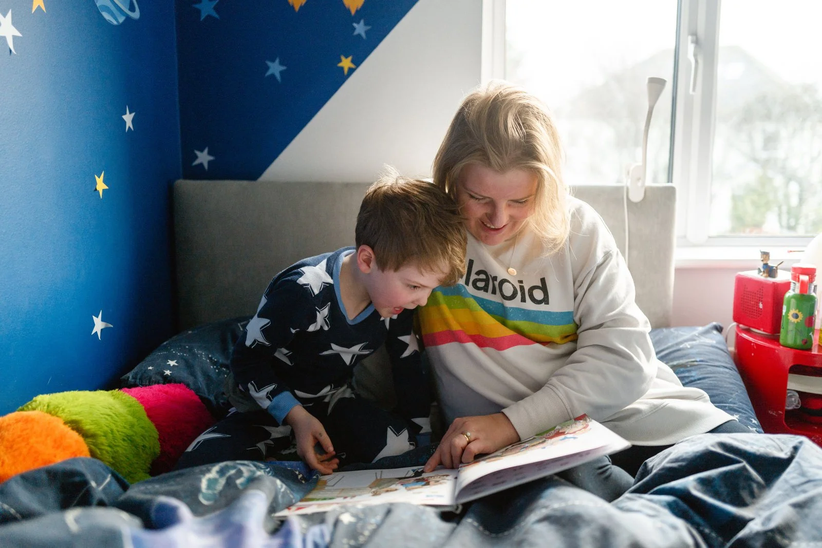 A woman and a young boy sitting on a bed, looking at a book together and smiling.