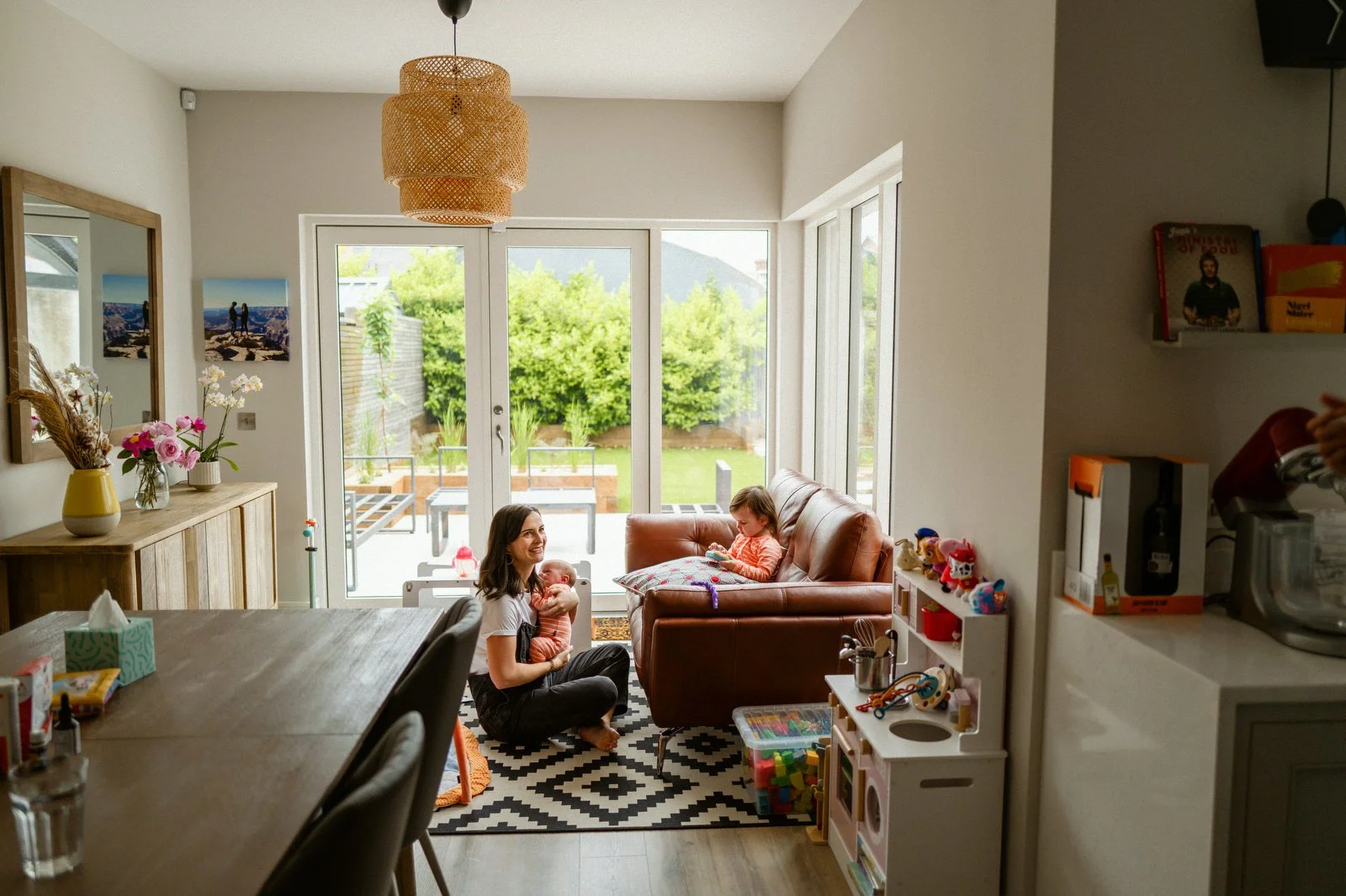 A woman sitting on the floor holding a baby, with another child sitting on a couch beside her in a bright living room with large glass doors leading to a green backyard.