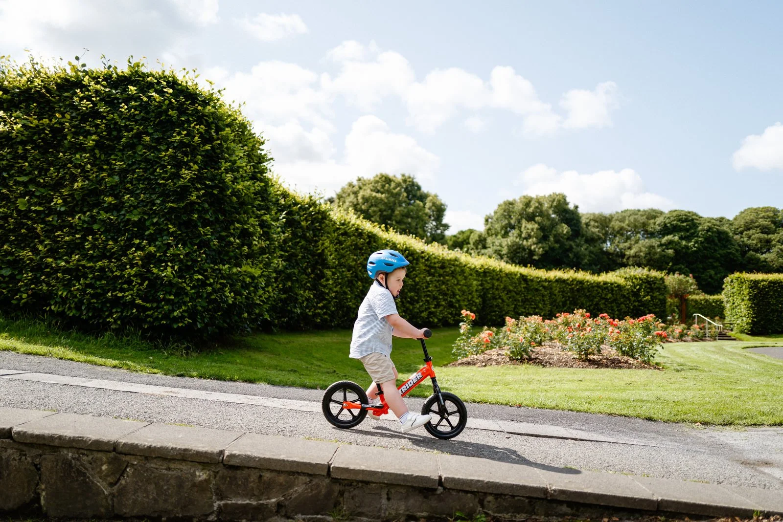 Young boy riding a red balance bike outdoors in a park with green bushes, flowering plants, and trees under a partly cloudy sky.