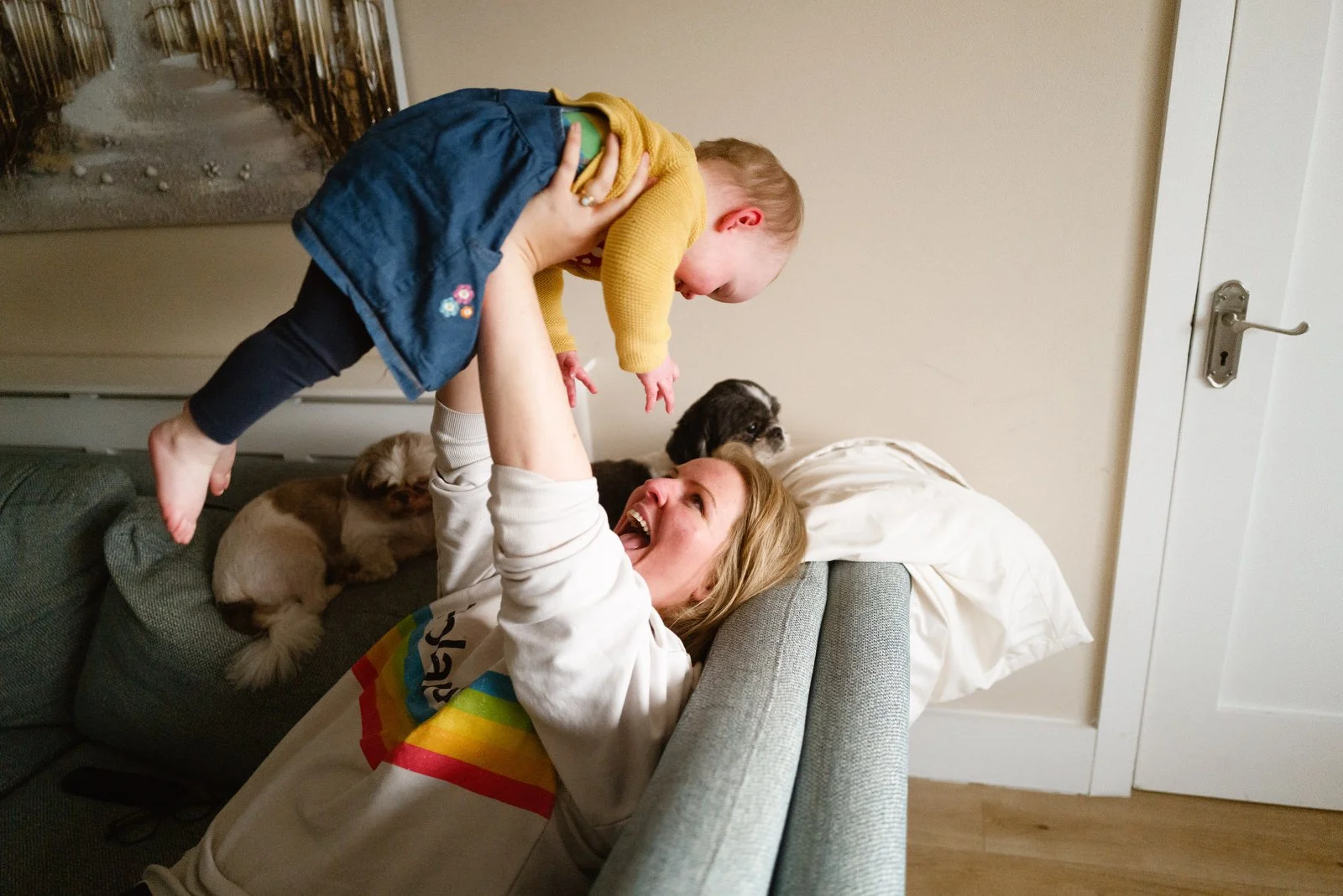 A woman lying on a sofa, smiling and playing with a young child who is holding onto her hands with their arms raised. The child is near the woman’s face. There are small dogs resting on the sofa behind them.