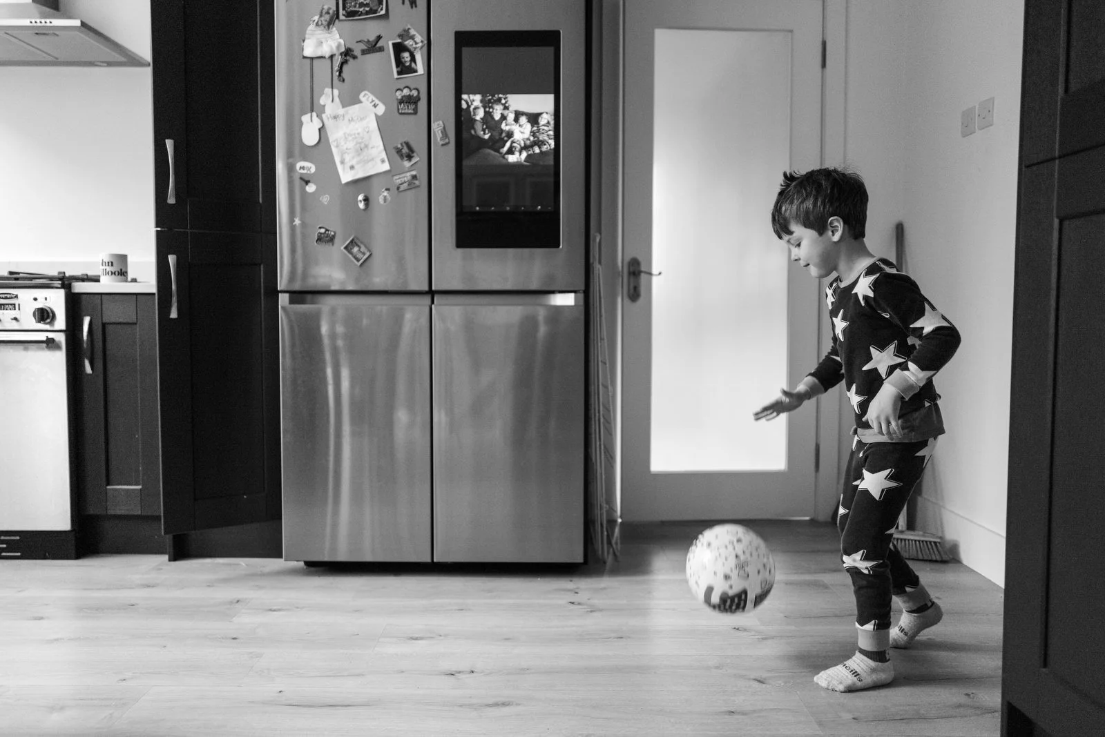 A young boy playing with a ball inside a kitchen, wearing pajamas with star patterns, near a refrigerator and a door.