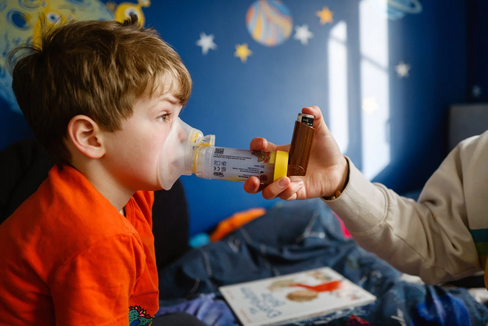 A young boy in an orange shirt is near a nebulizer mask pressed to his face, with an adult holding the nebulizer medication bottle. The background shows a blue wall with decorations and a window letting in natural light.