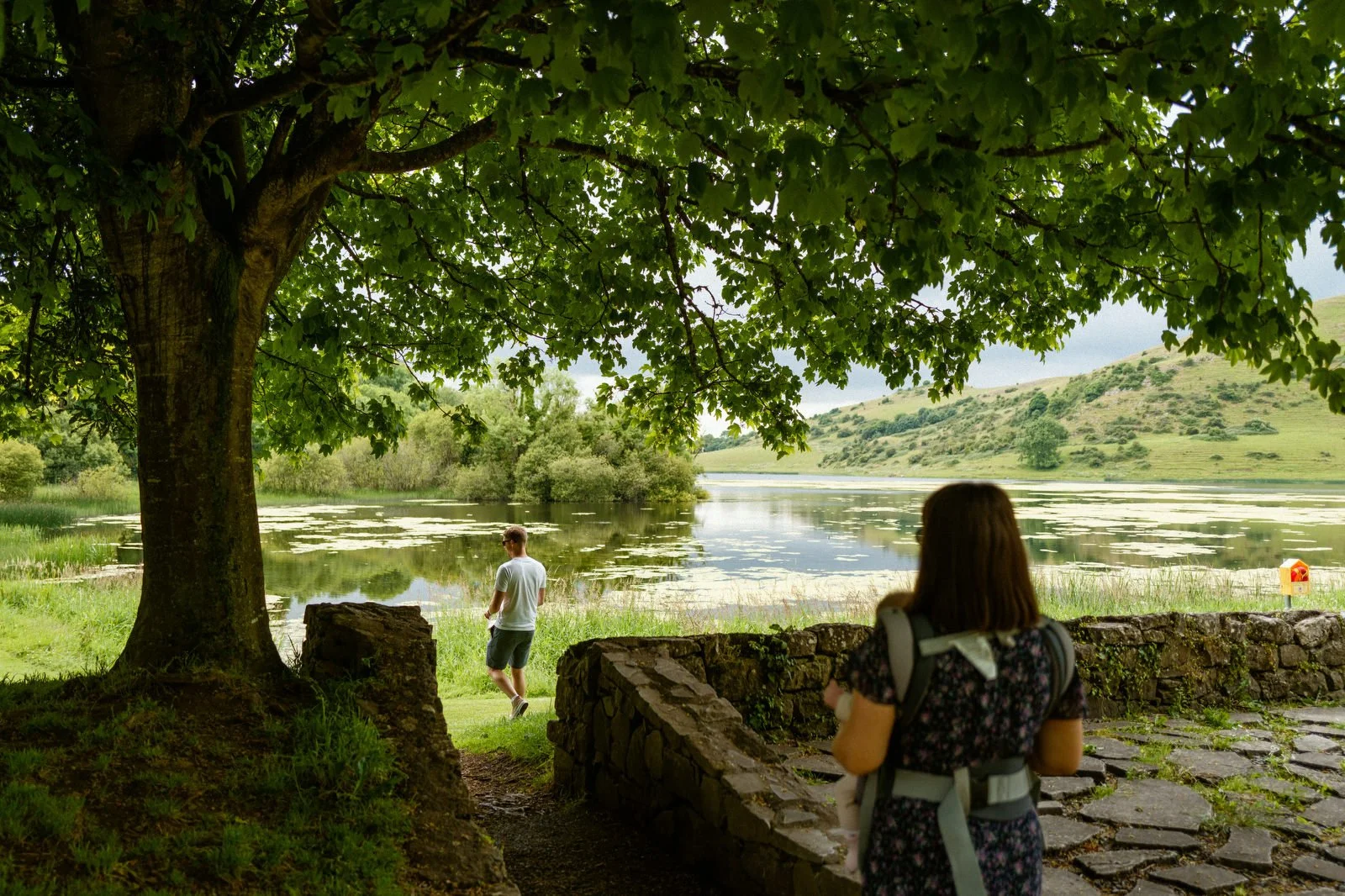 A woman with a backpack standing on a stone path by a lake, observing a man walking by the water under a large leafy tree.