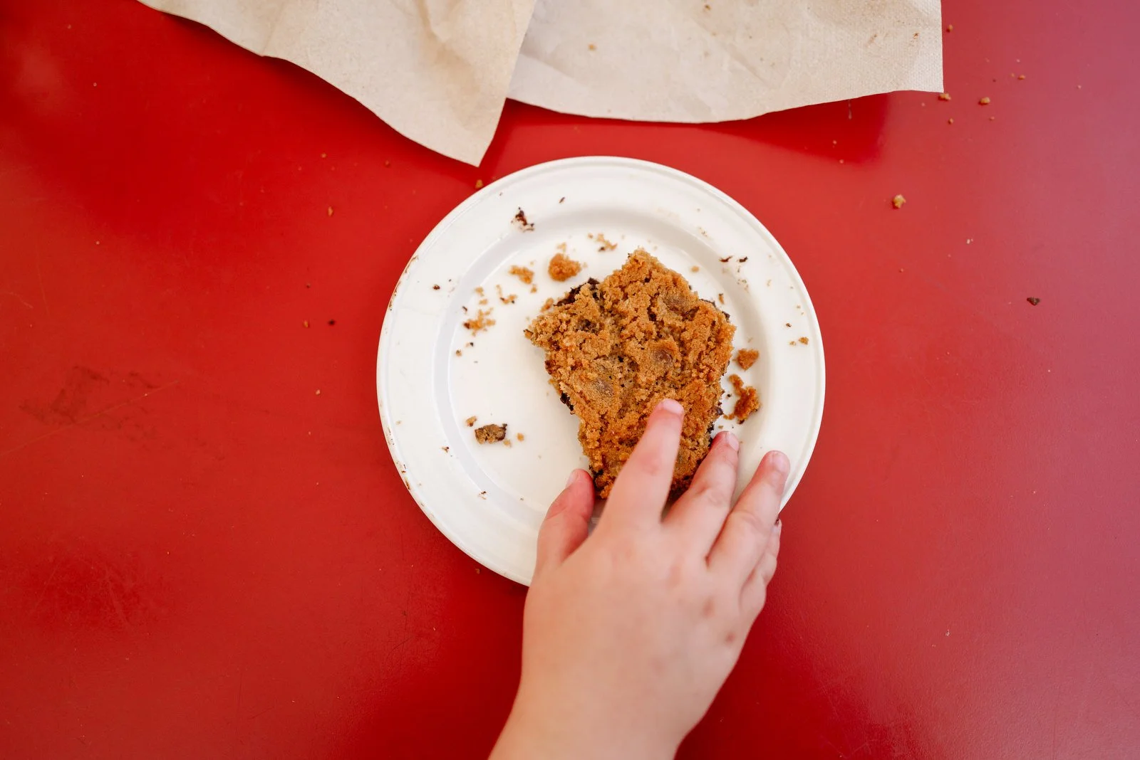 Child's hand reaching for a piece of cookie on a white paper plate, on a red table with crumbs around.