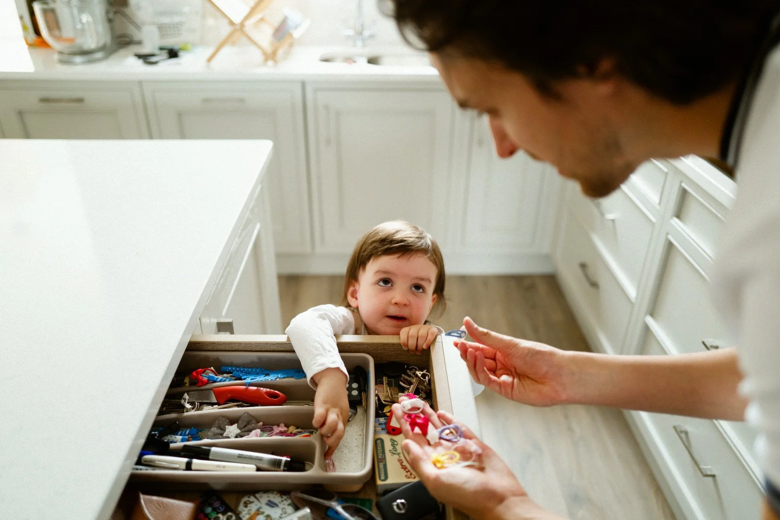 A young child reaching into a drawer filled with various small items and toys, while an adult shows or hands the child a small object in a kitchen