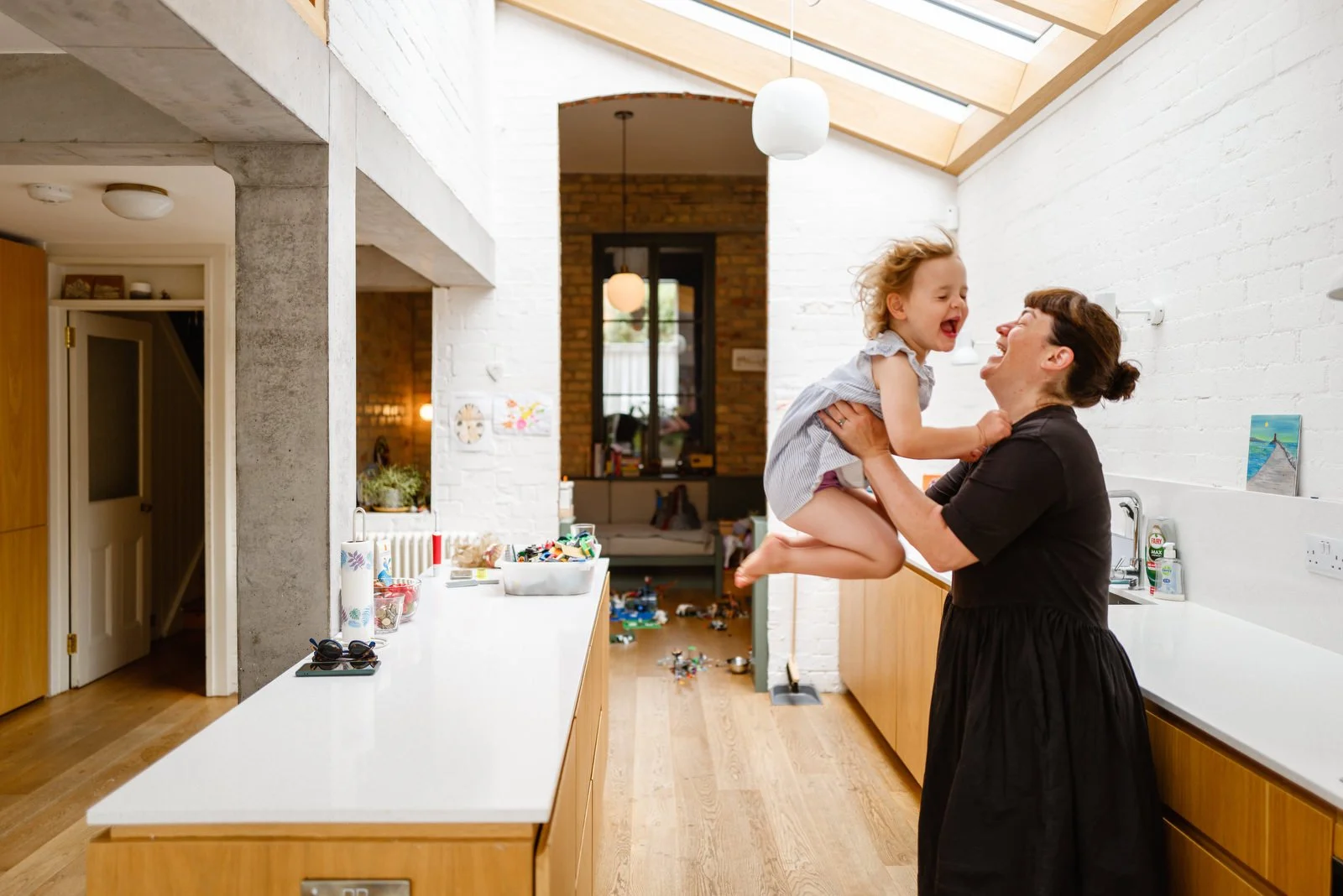 A woman lifting a young girl in a kitchen, both smiling and enjoying a playful moment. The kitchen has white brick walls, wooden cabinets, and a skylight ceiling.