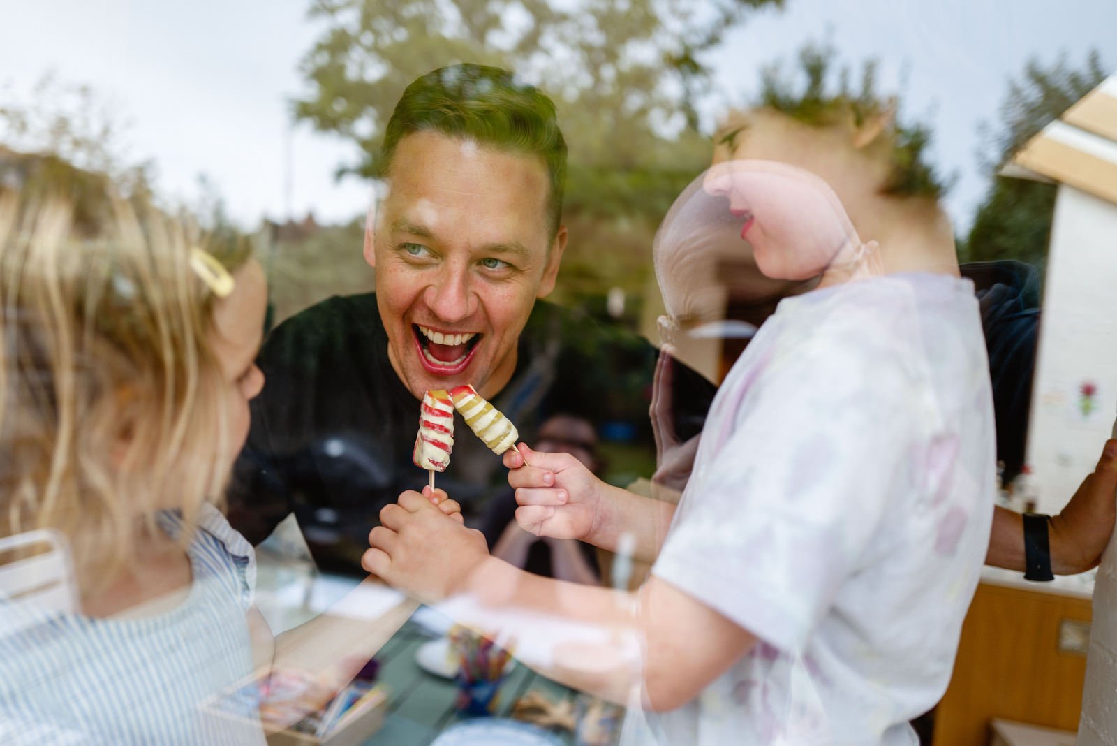 A man smiling joyfully as he shares ice cream popsicles with two young girls through a glass window during a celebration.