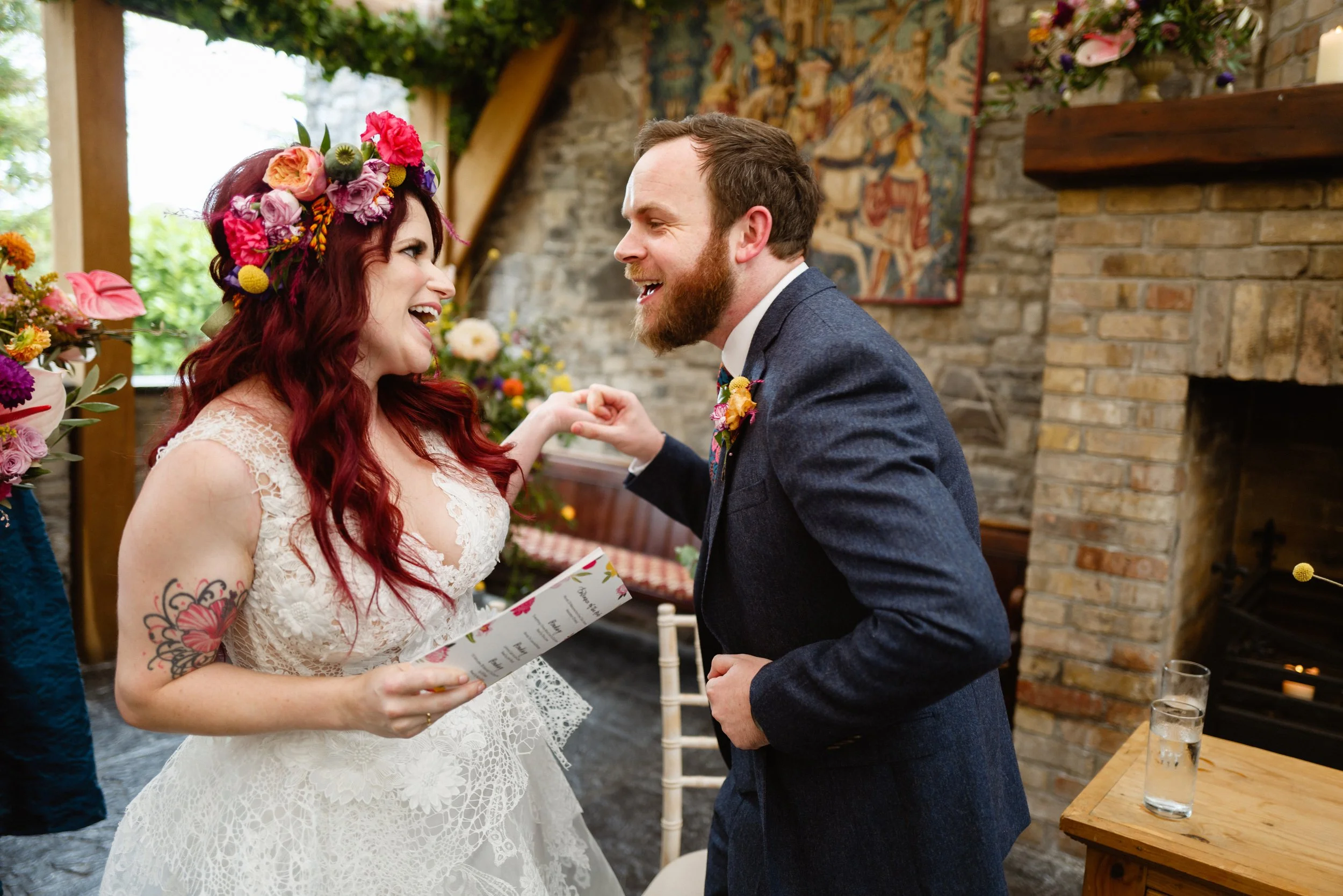A couple at a wedding, the woman with long red hair, a flower crown, a lace dress, and holding a paper, and the man in a dark suit and colorful tie, with a background of a brick fireplace, floral arrangements, and a tapestry.