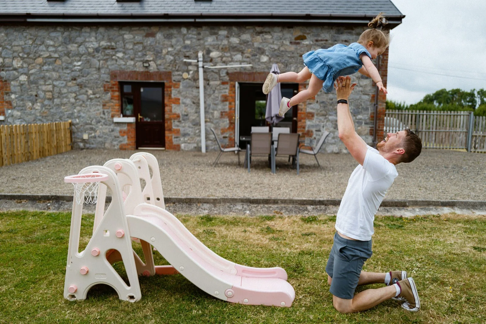 A man kneeling on the grass and holding a young girl up in the air outside, with a small pink and white plastic slide and a stone house with a patio in the background.