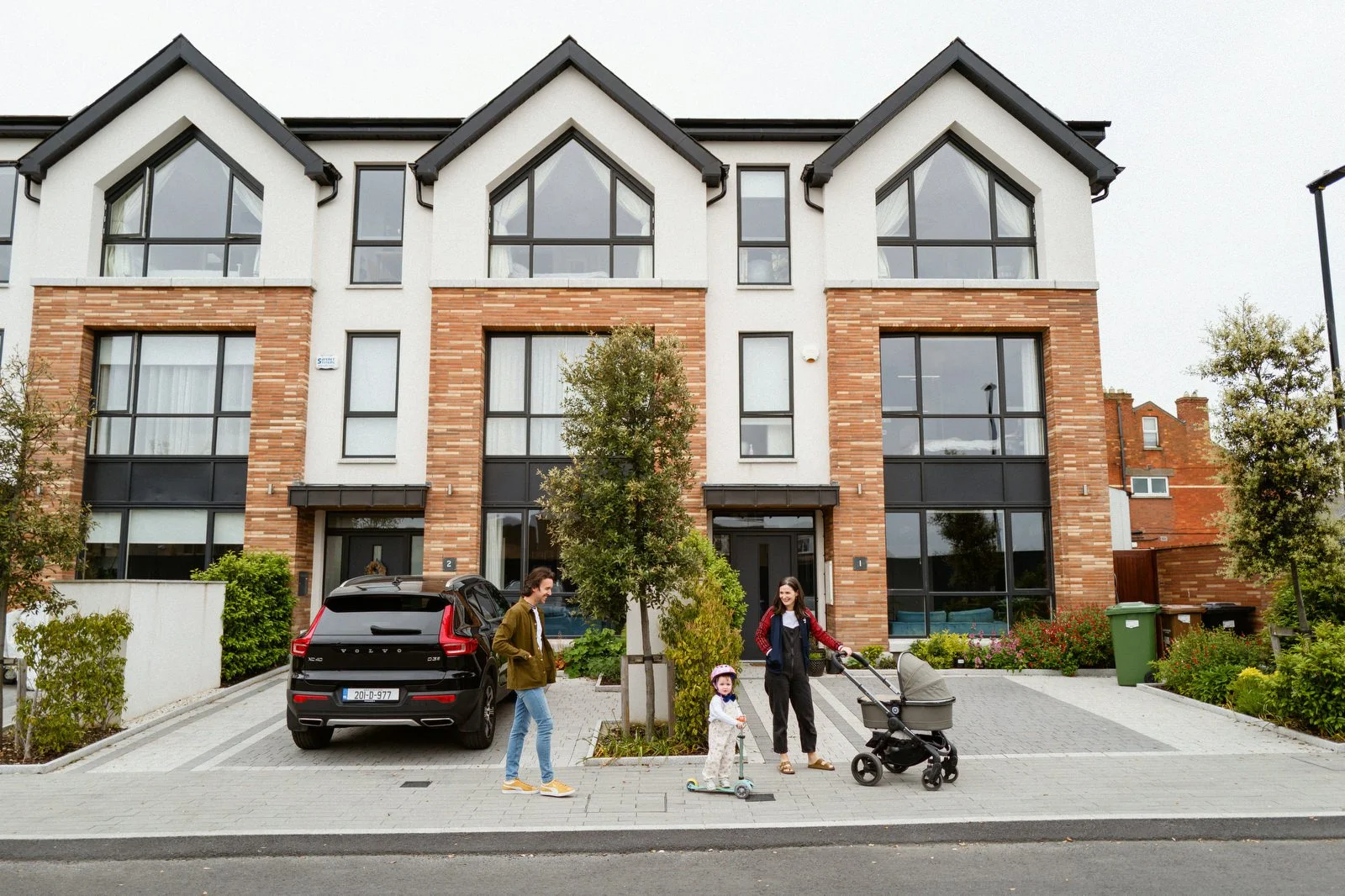 Three people, one child on a scooter and a woman with a stroller, stand on a sidewalk in front of a modern residential building with brick and white exterior, large windows, and a parked car.