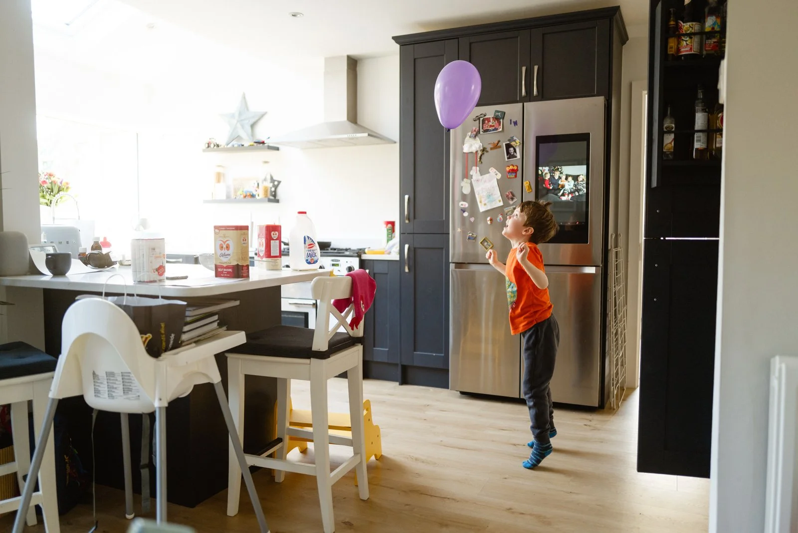 A young boy is playing with a purple balloon in a kitchen, standing next to a stainless steel refrigerator decorated with various magnets and photos. The kitchen has dark cabinets, a white countertop, and several food items and containers on the coun