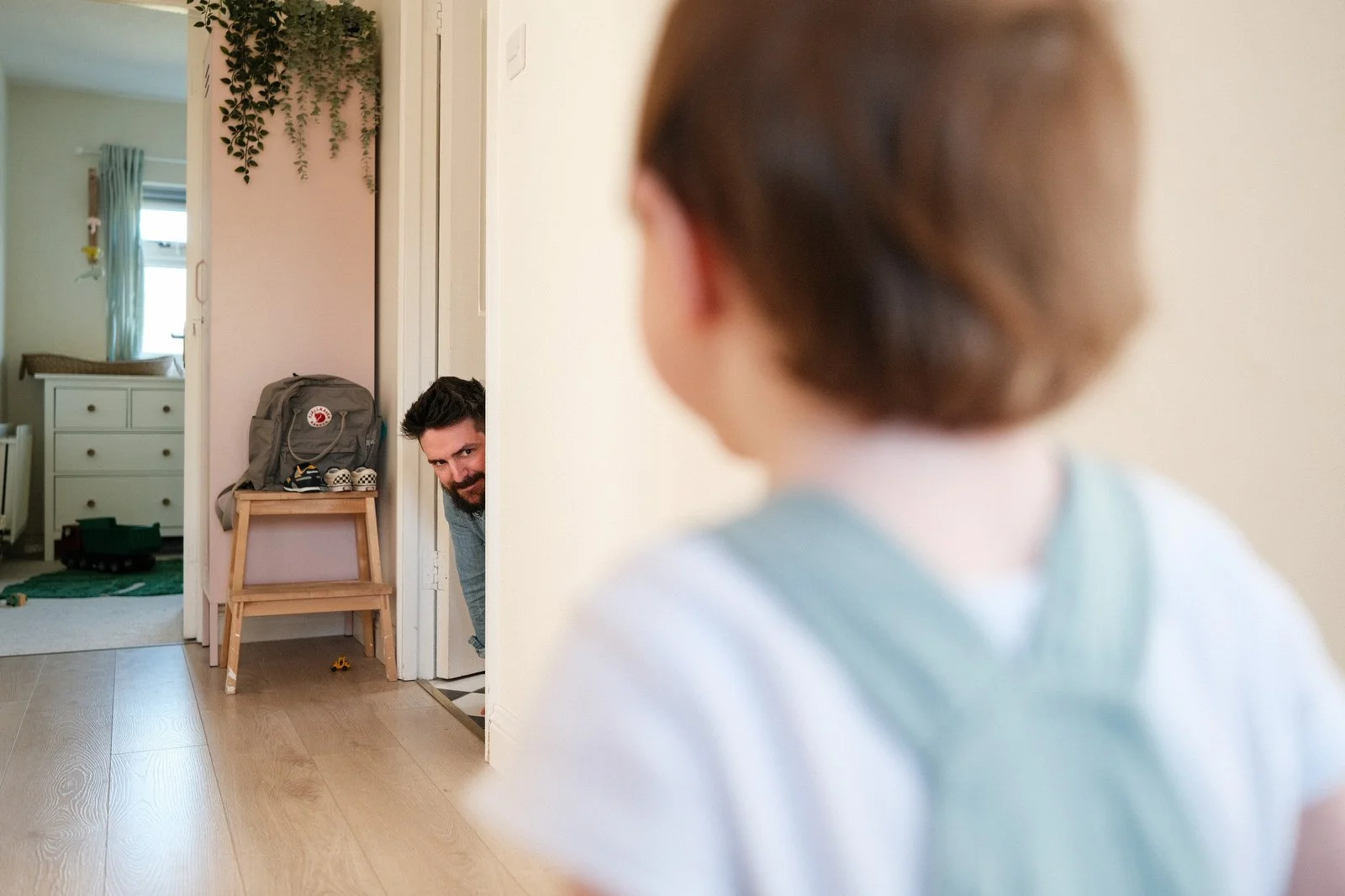 A young girl looking at a man peeking out from behind a doorframe in a home interior.