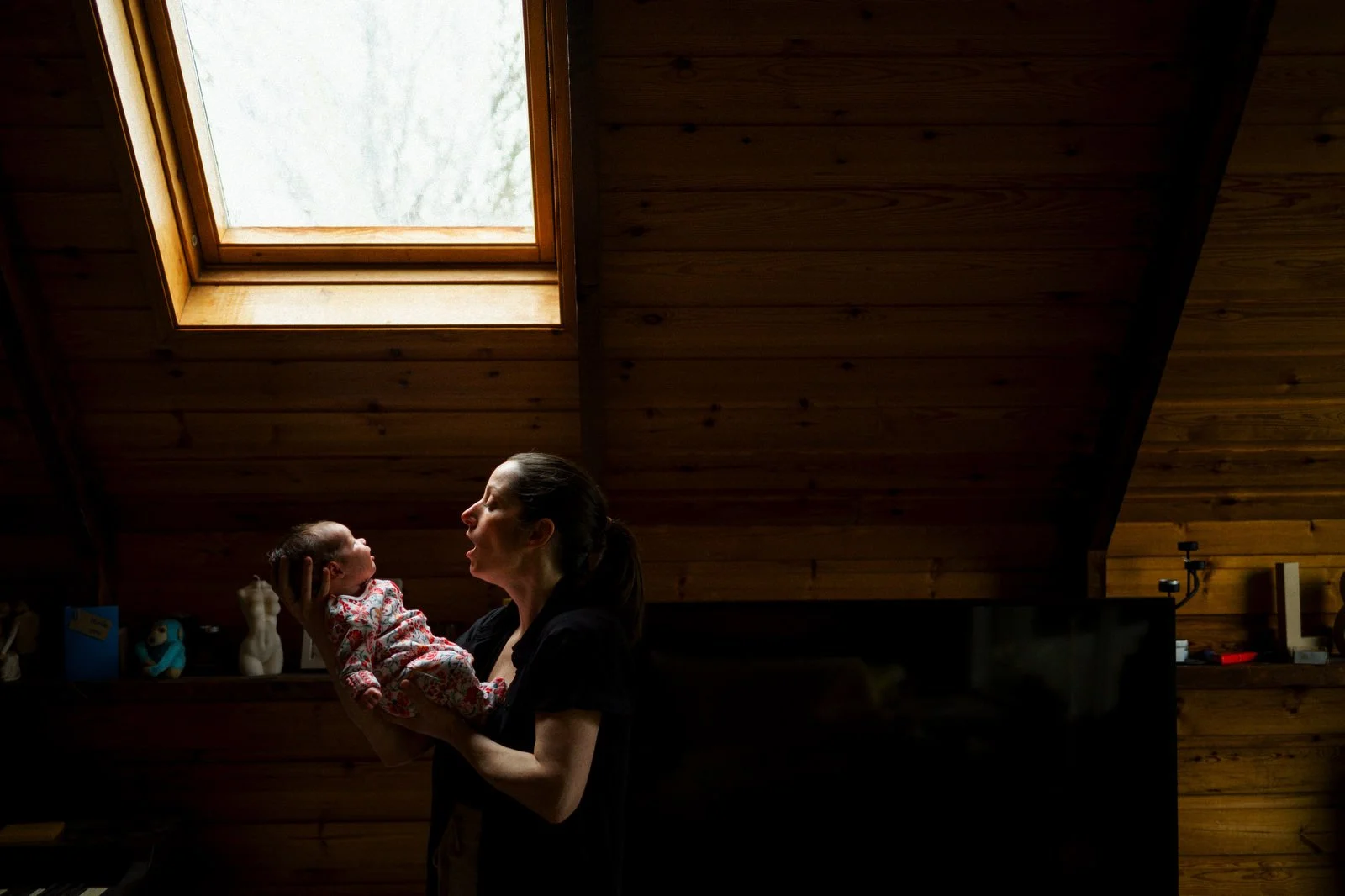 A woman is holding a newborn baby inside a room with wooden walls, near a skylight window in the ceiling.