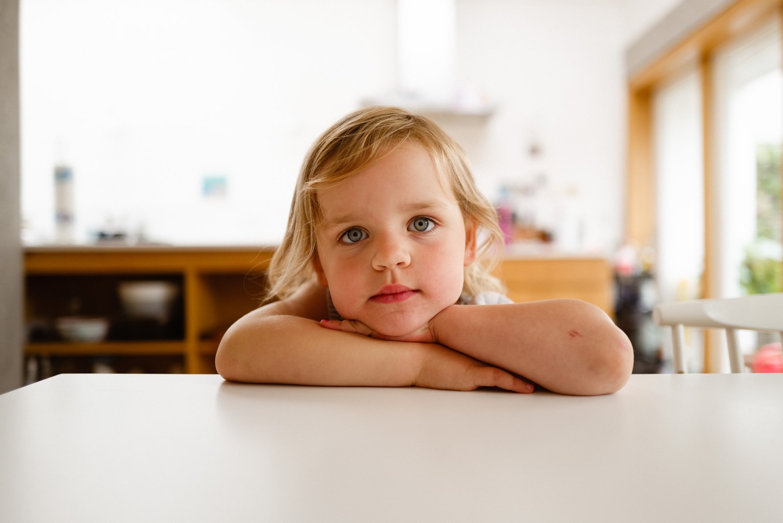 A young girl with blonde hair and blue eyes resting her chin on her folded arms on a white table in a bright, modern kitchen.