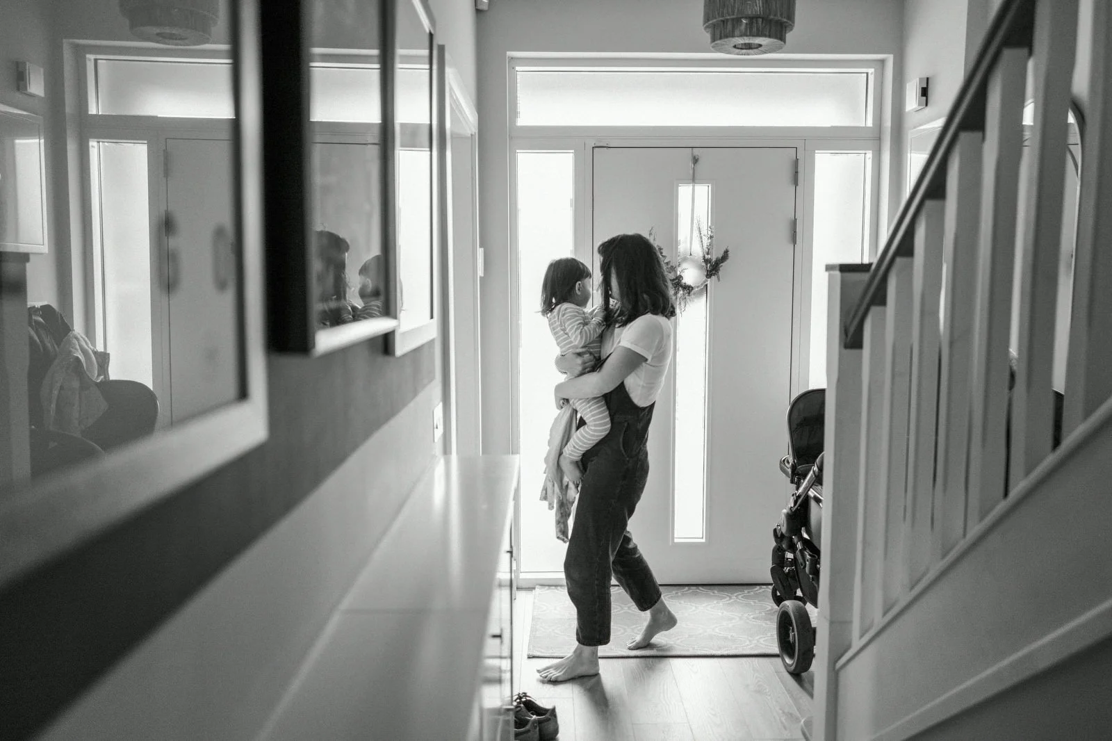 A woman holding a young girl near the front door of a house, with a stroller and stairs visible in the background, in a black-and-white photo.