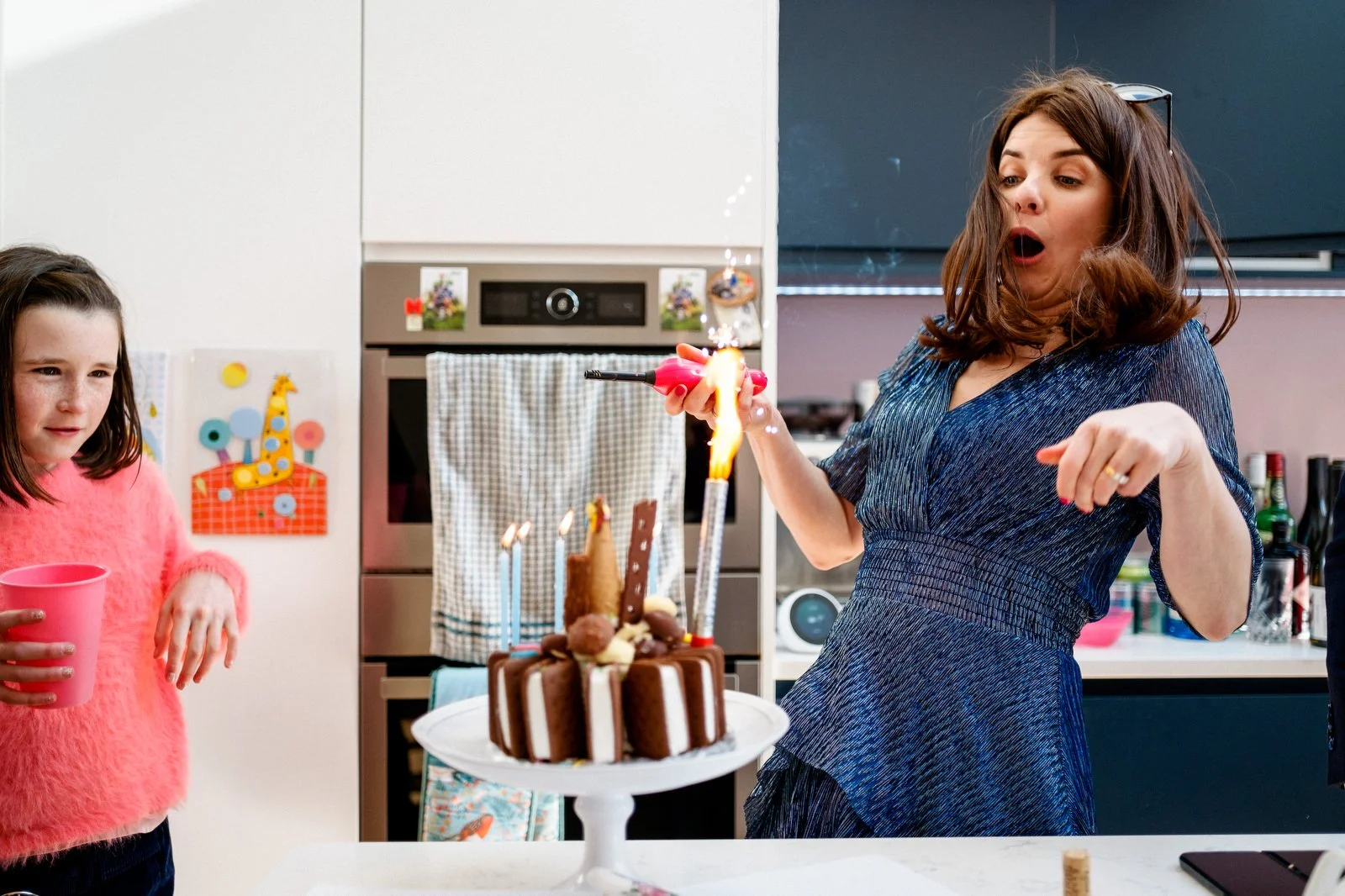 A woman is surprised as she lights candles on a birthday cake. Another girl is watching with a drink in hand in a kitchen.