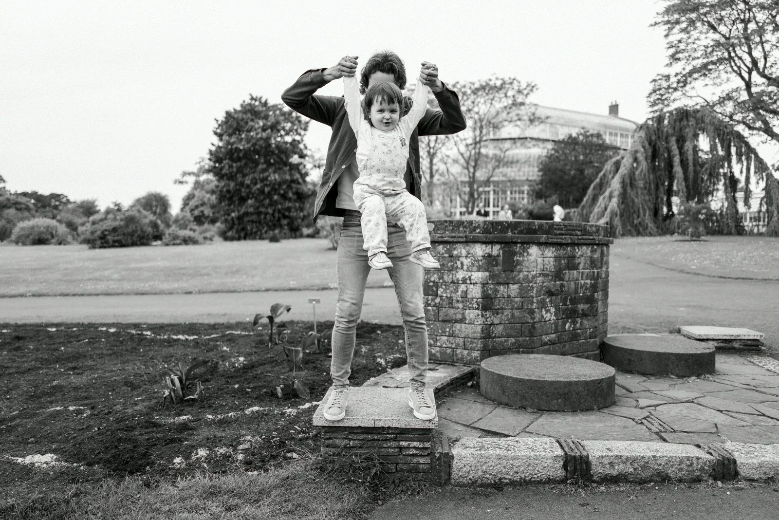 A woman holding a young girl up in the air in a park with trees and a large building in the background.