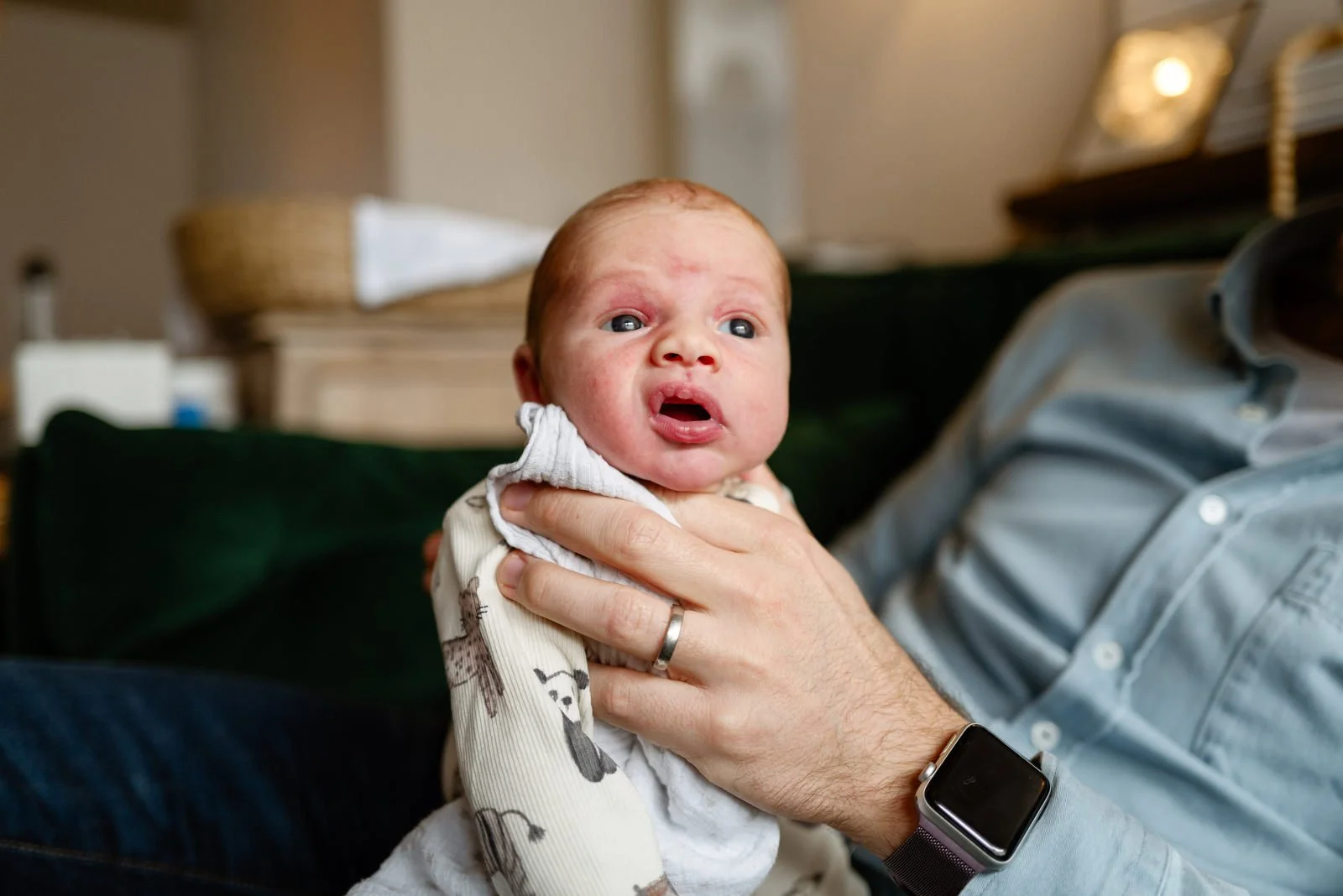 A baby with red hair and blue eyes, being held by an adult wearing a gray shirt and a smartwatch, inside a house.