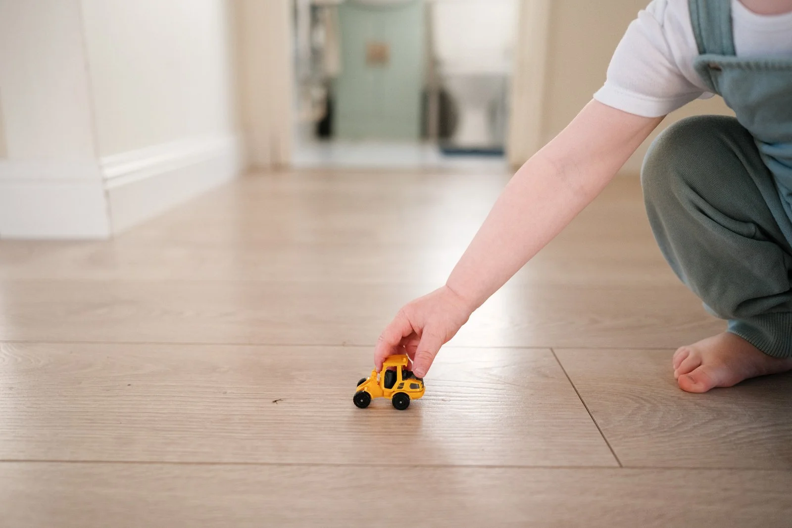 A child playing with a yellow toy bulldozer on a light wooden floor in a house.