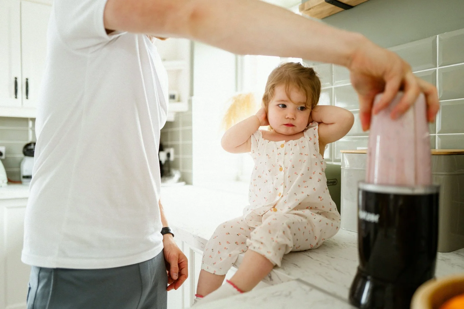 A young girl sits on a kitchen counter with her hands over her ears, looking at a man holding a pink object, possibly a blender, on a kitchen counter.