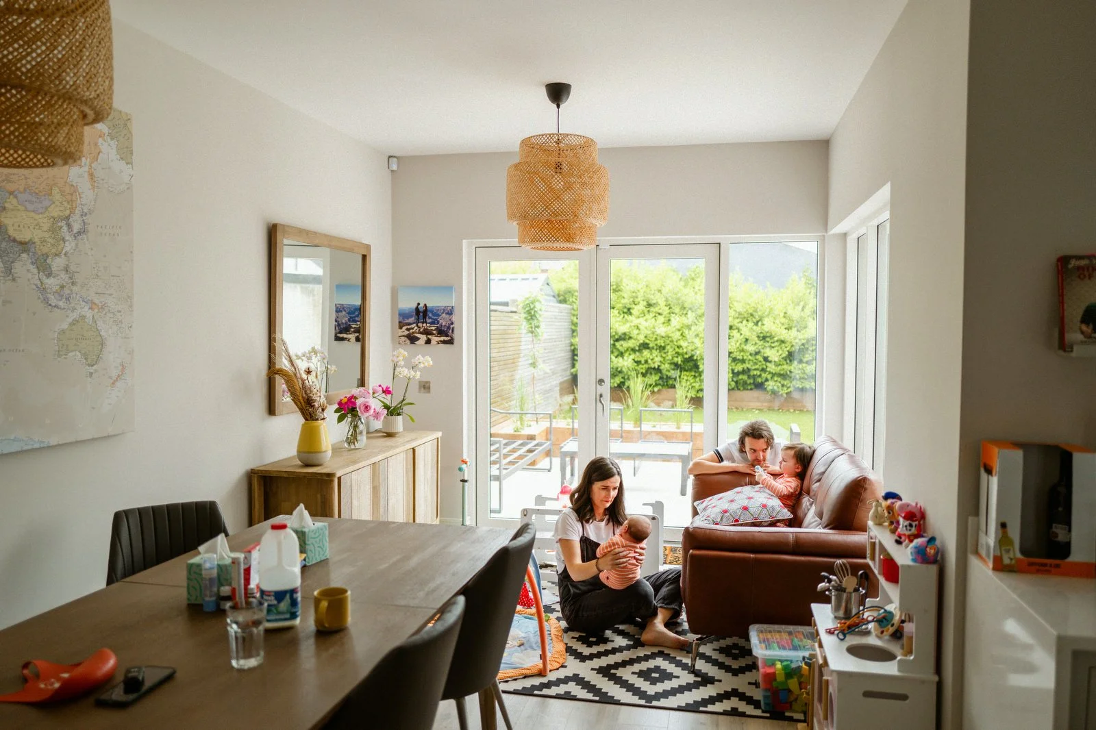 A living room with a family engaging in activities: a woman holding a baby, a girl sitting on the floor, and a man lying on a sofa. Large glass doors lead to a backyard with greenery.