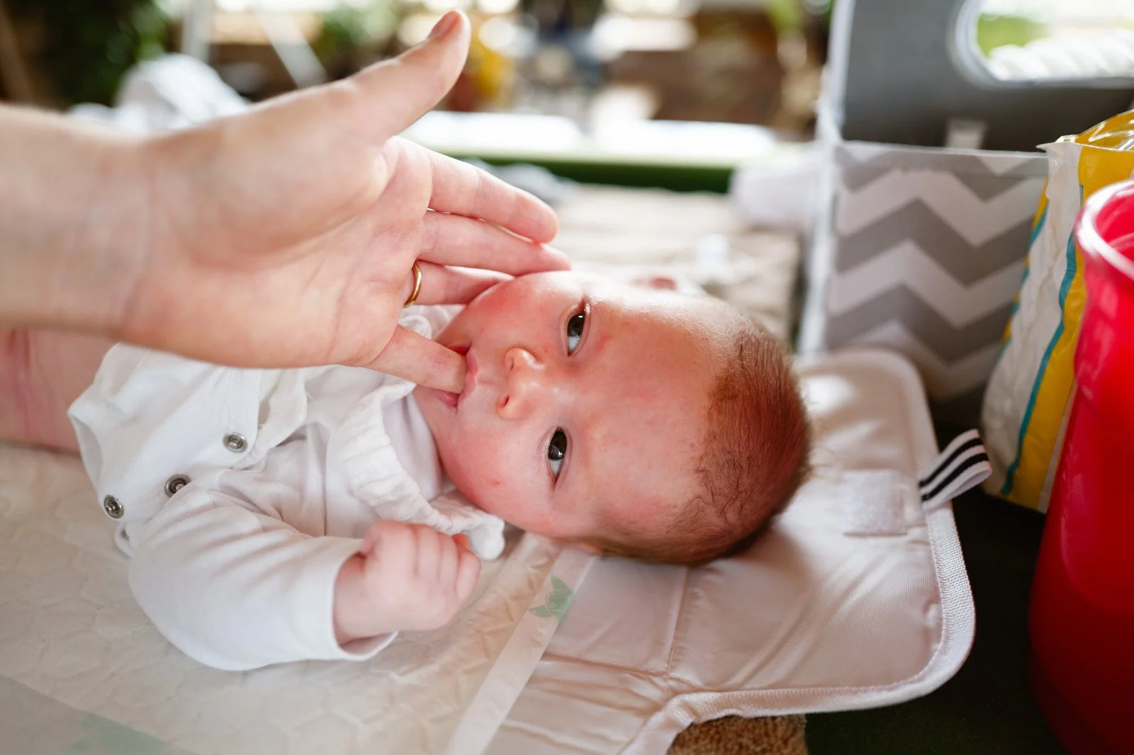 A baby lying on a changing pad, receiving a finger-fed thumb in mouth from an adult hand