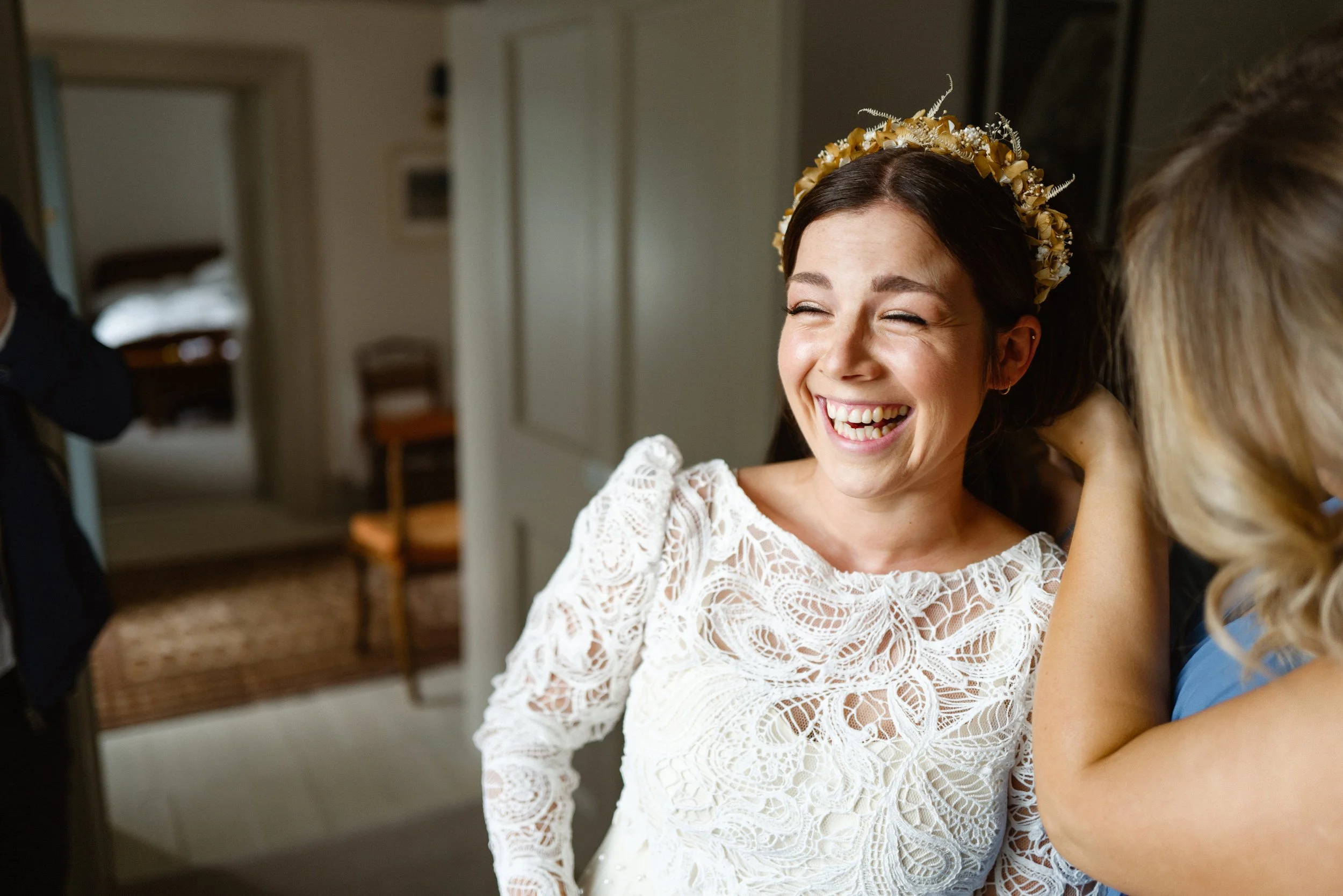 A woman in a white lace dress with a floral headband smiling and laughing, possibly a bride getting ready for her wedding.