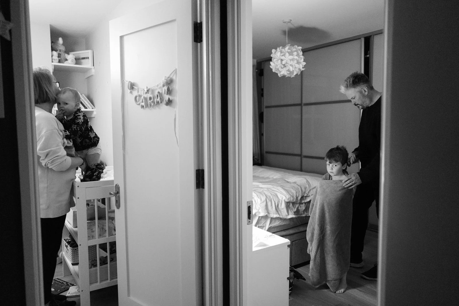 A view of two bedrooms, one with a woman holding a child and the other with two men grooming a child on a bed, as seen through a doorway.