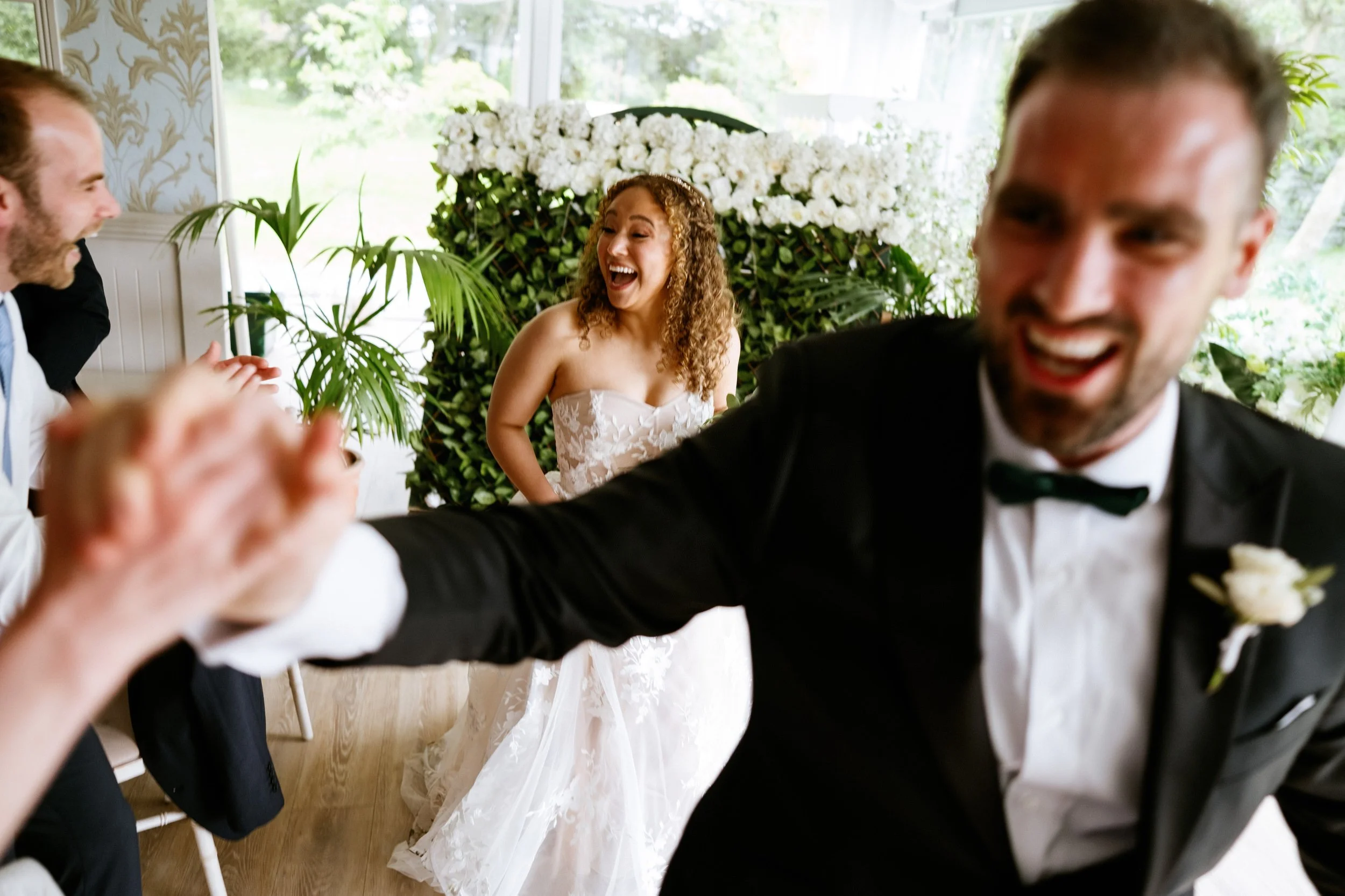 Wedding celebration with people smiling and dancing, bride in a white dress, groom in a black tuxedo, and floral decorations in the background.