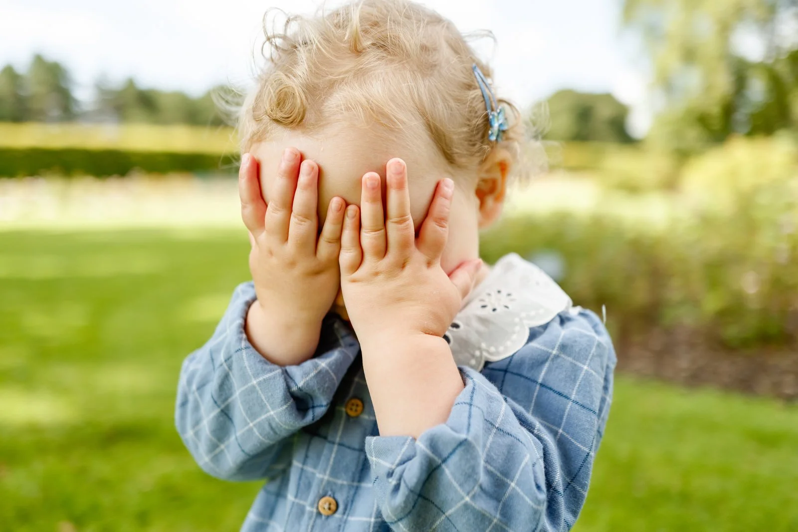 Child with curly blond hair, wearing a blue checkered shirt with white lace collar, covering her face with her hands outdoors in a park with green grass and trees.