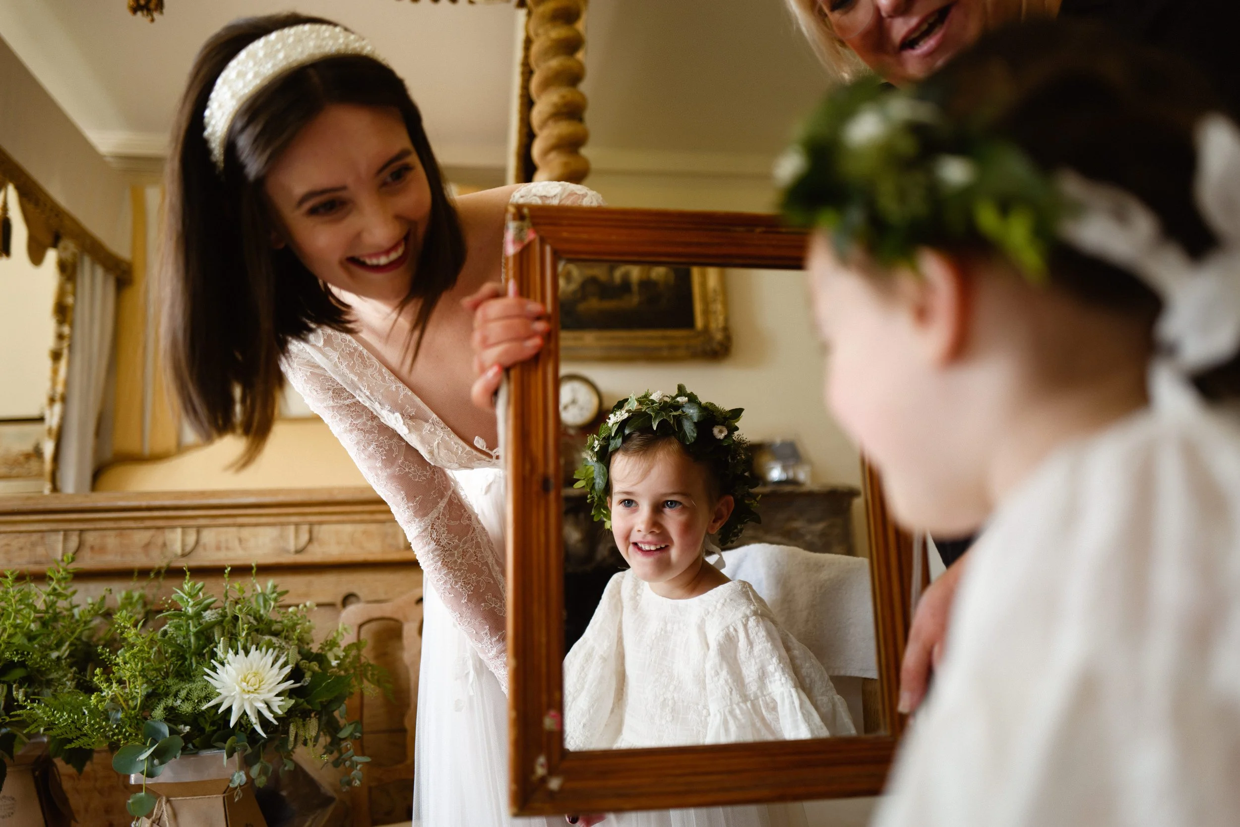 A young girl dressed in white with a floral crown is looking into a mirror, smiling. Two women, one with dark hair and a white dress with lace sleeves, and another with blonde hair are admiring her reflection during a wedding or special occasion in a