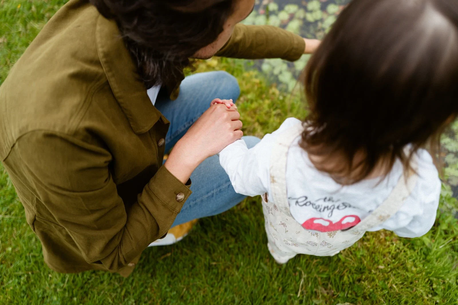 An adult holding a child's hand with both sitting on grass.