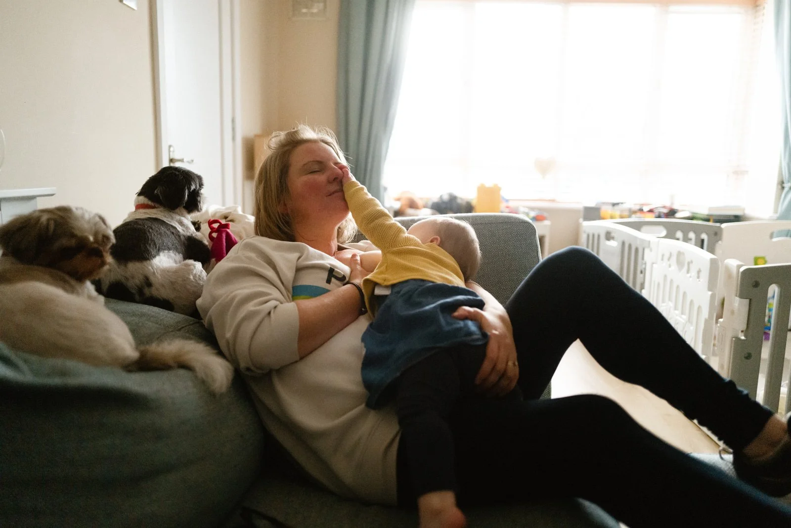 A woman sitting on a couch with two puppies beside her and a young child leaning on her chest, touching her nose. She appears to be relaxed and happy inside a living room with bright window light.