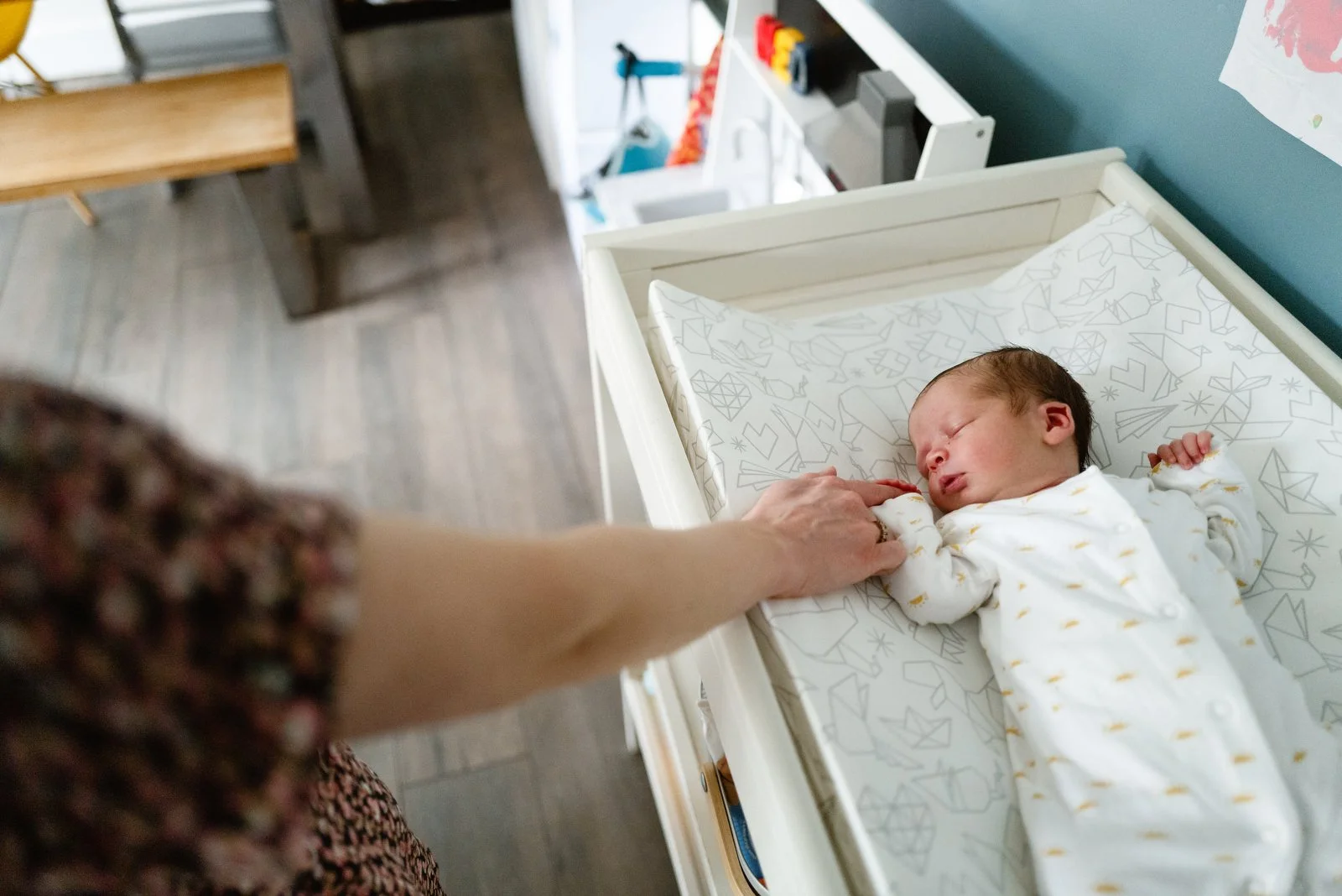 A newborn baby lying in a crib, with an adult's hand gently holding the baby's hand. The baby is sleeping, wearing a white onesie with yellow patterns. The crib has a white frame and a patterned mattress.