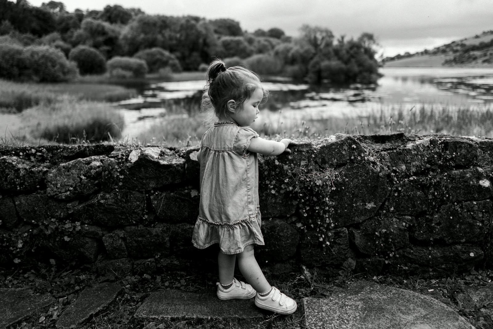 A young girl with pigtails in a dress and sneakers leaning on a stone wall, looking out over a lake with trees and hills in the background.