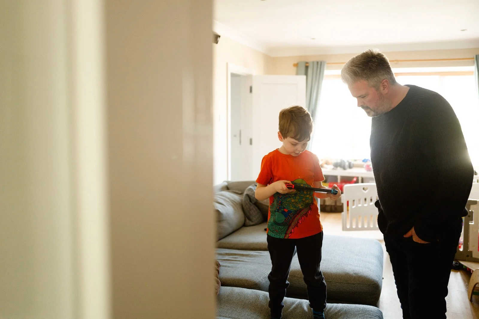 A boy with a tablet standing on a couch, looking at the screen, with an older man with gray hair and beard standing beside him in a well-lit living room.