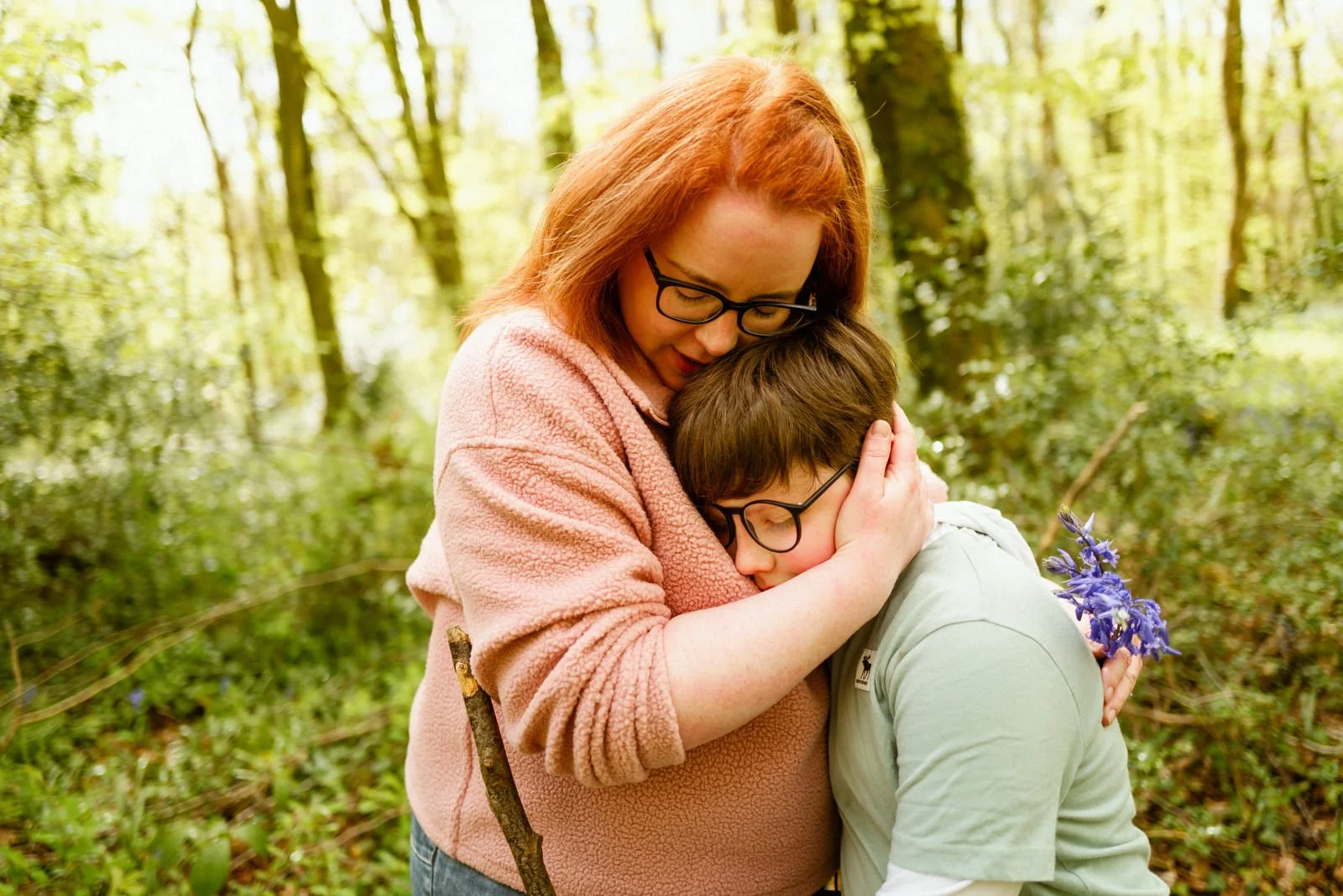 A woman with red hair and glasses hugs a young boy with glasses, holding a purple flower, in a forested area.