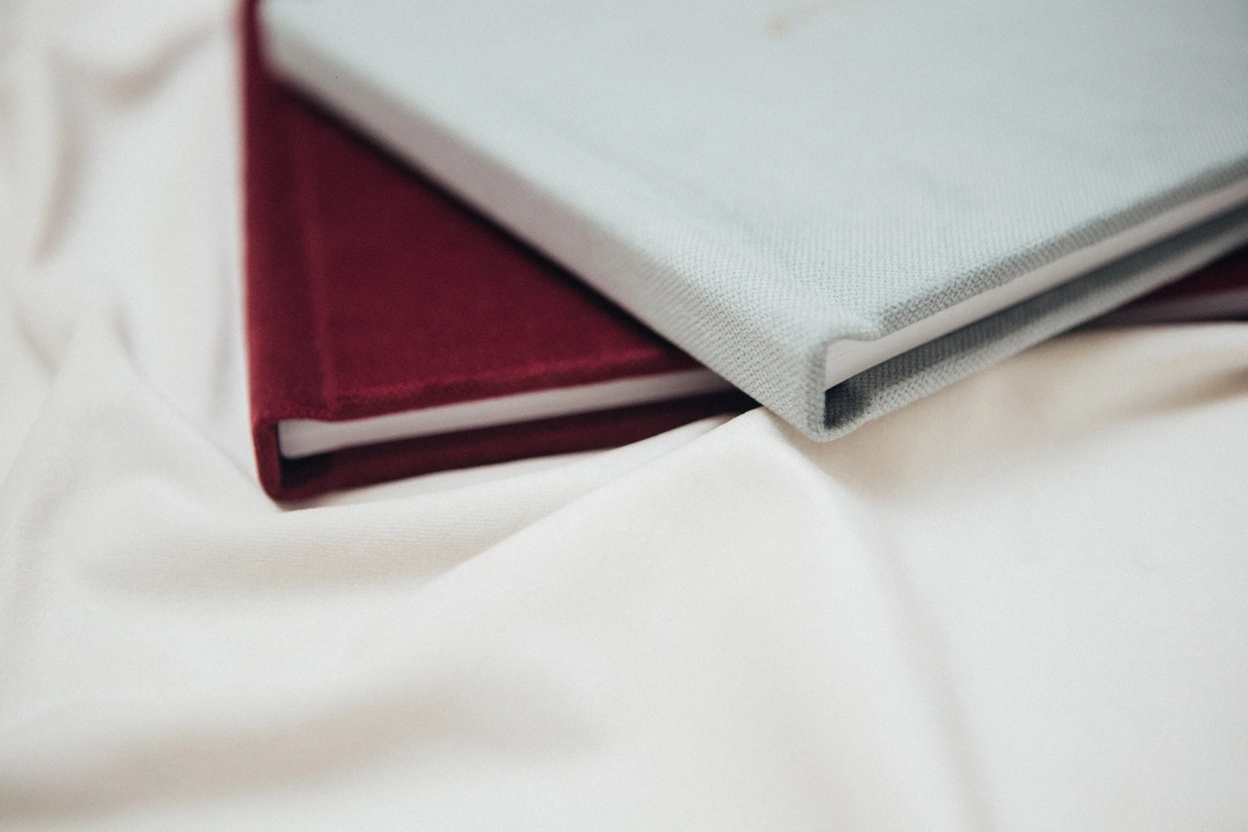 Two closed hardcover books, one with a red cover and one with a gray cover, resting on a cream-colored fabric surface.
