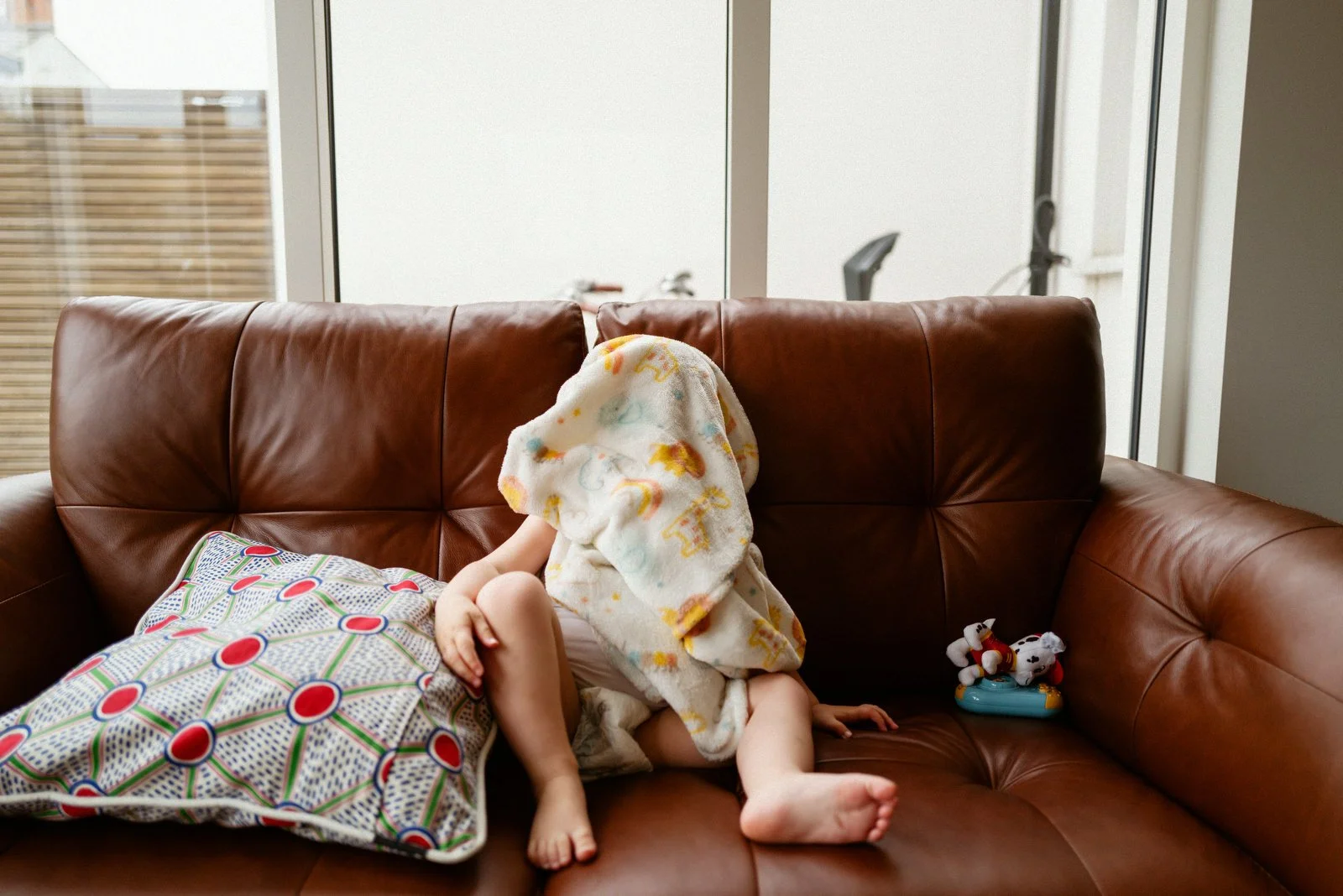 Child sitting on a brown leather couch with a blanket over their head, surrounded by pillows and plush toys.