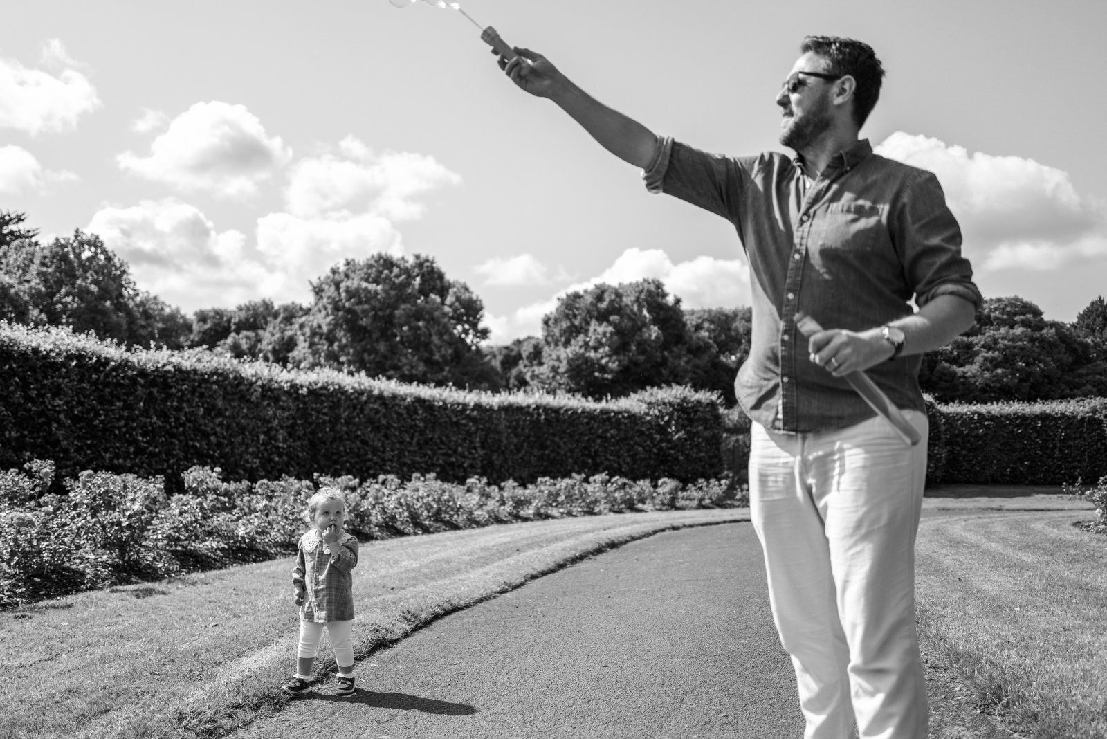 A man with glasses and a beard is holding a spray bottle and a spray gun outdoors on a curved pathway, with a young girl standing at a distance and looking at him, surrounded by trees and bushes on a partly cloudy day.