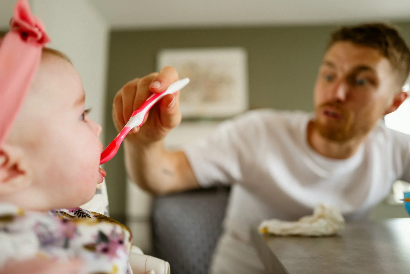 A young girl with a pink bow in her hair being fed a piece of cake with a pink and white striped spoon by a man, possibly her father, at a table indoors.