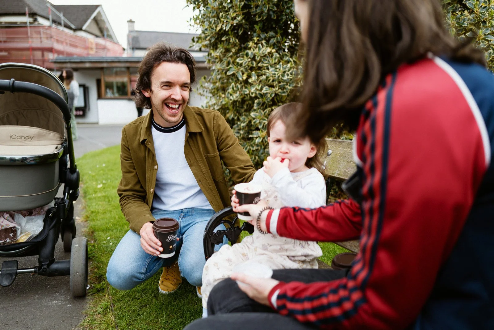 A young man sitting on a bench outdoors, laughing, with a young girl and a woman, possibly his family, drinking coffee and enjoying time together.