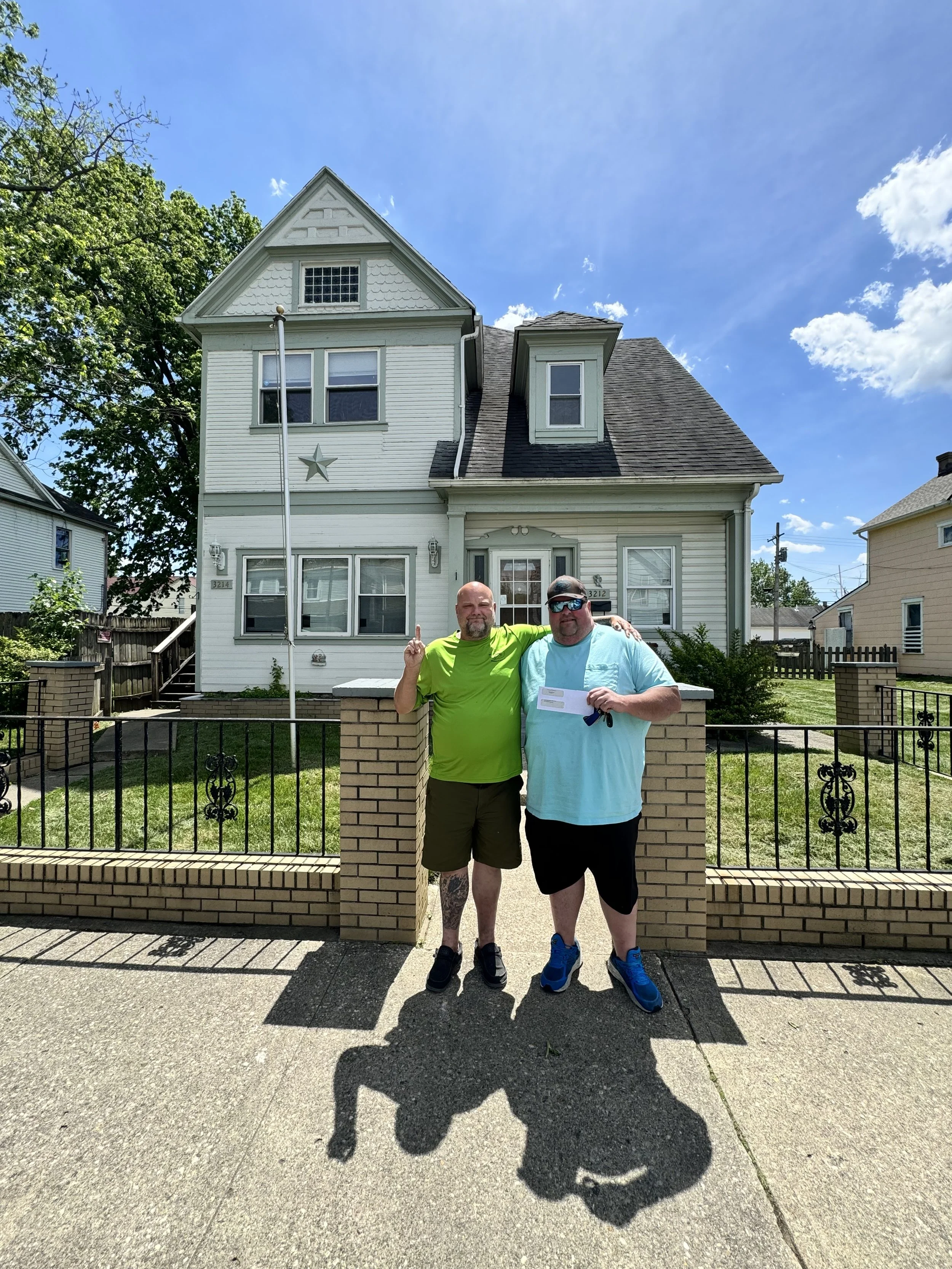 Two men standing in front of a two-story house with a peaked roof on a sunny day, with one man holding a piece of paper.