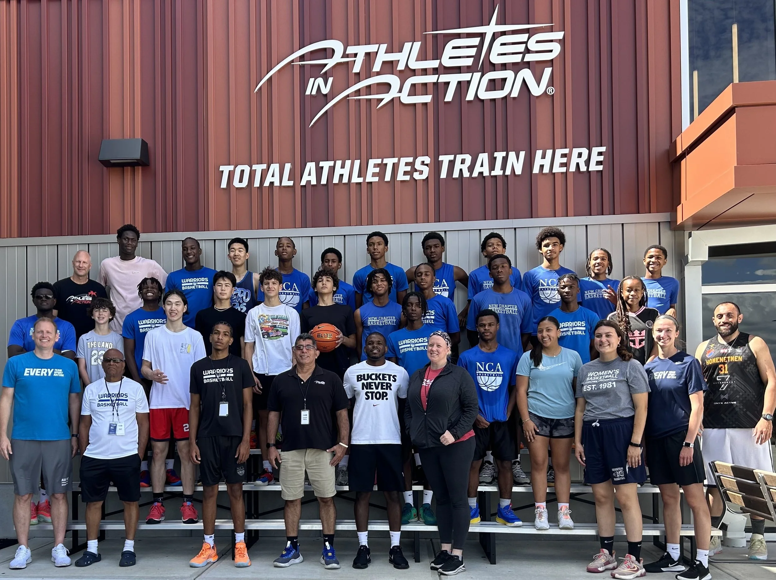A group of people, including athletes and coaches, stand on bleachers in front of an 'Athletes in Action' sign. They are wearing various sports attire, with some wearing t-shirts representing basketball teams. The building's sign reads 'Total Athletes Train Here.'