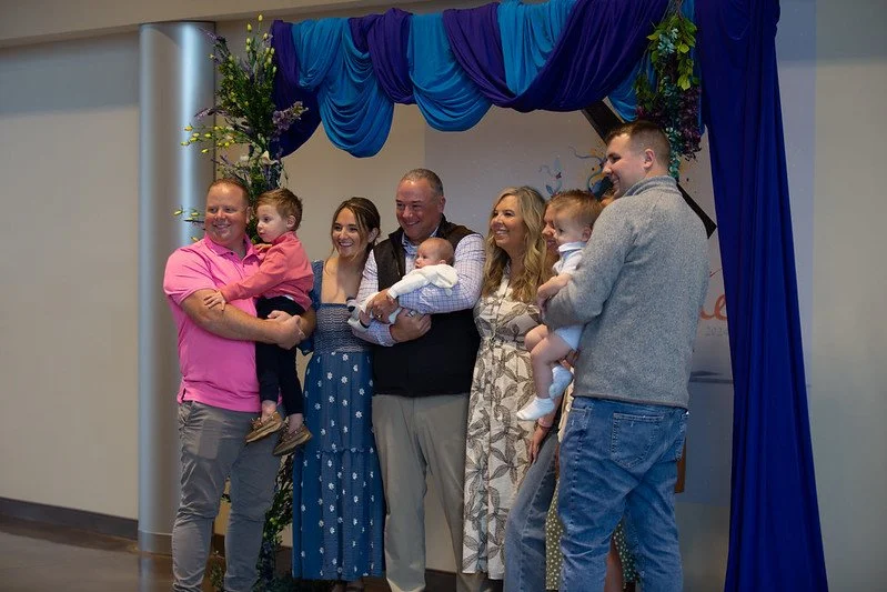 A group of adults and children pose together at an event with purple and blue drapery and floral decorations in the background.
