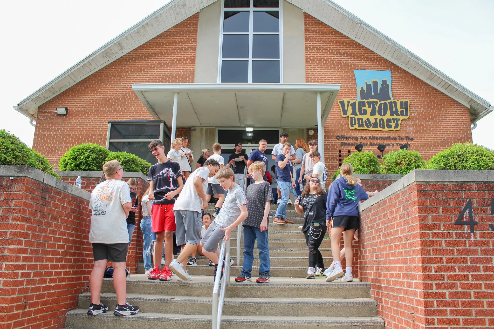 Group of people standing on stairs outside a brick building with "Victory Project" sign